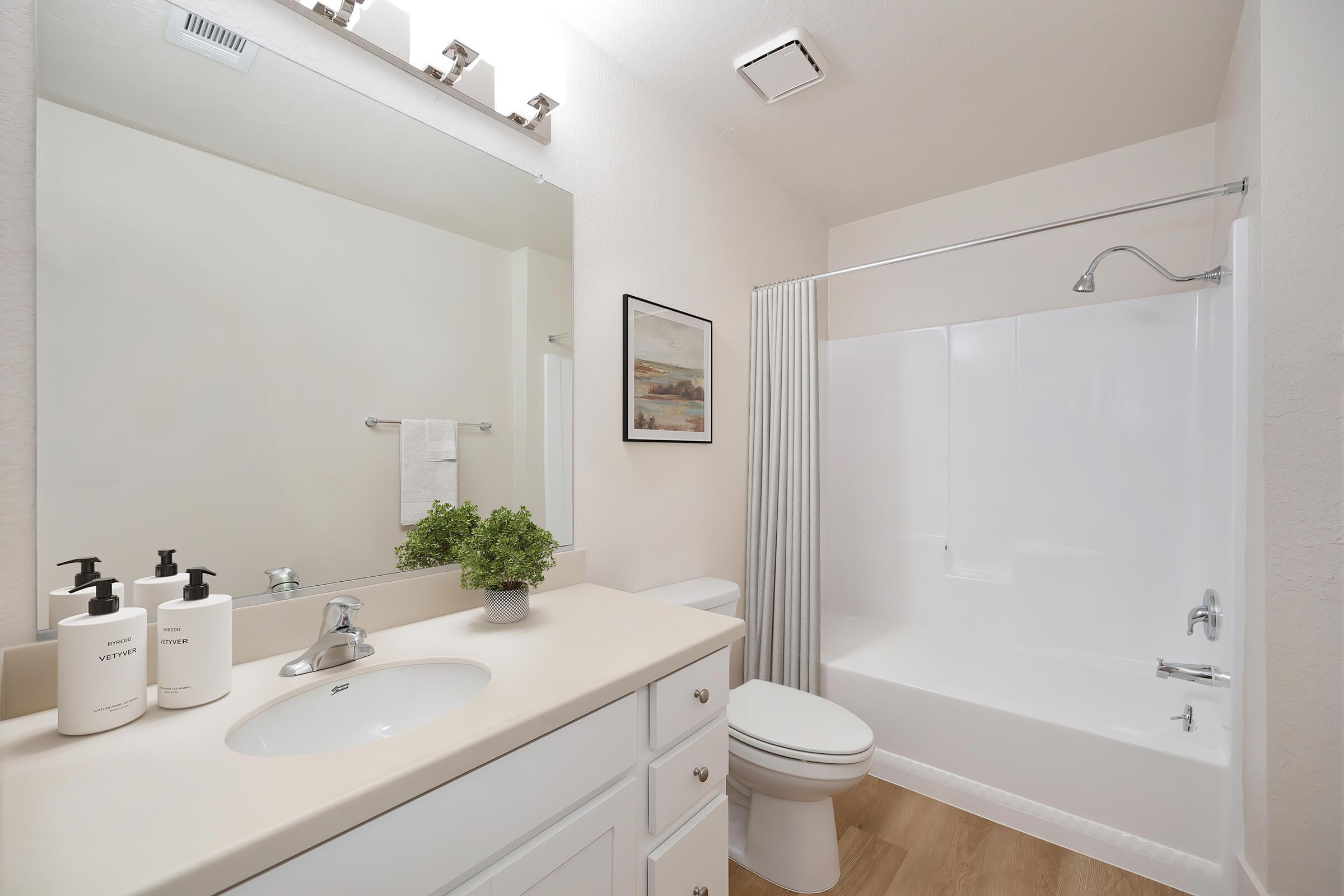 A bright and clean bathroom featuring a white vanity with a sink, three decorative bottles, a framed artwork on the wall, a shower curtain, and a bathtub. The space has light-colored walls and a neutral wooden floor, creating a modern and inviting atmosphere.