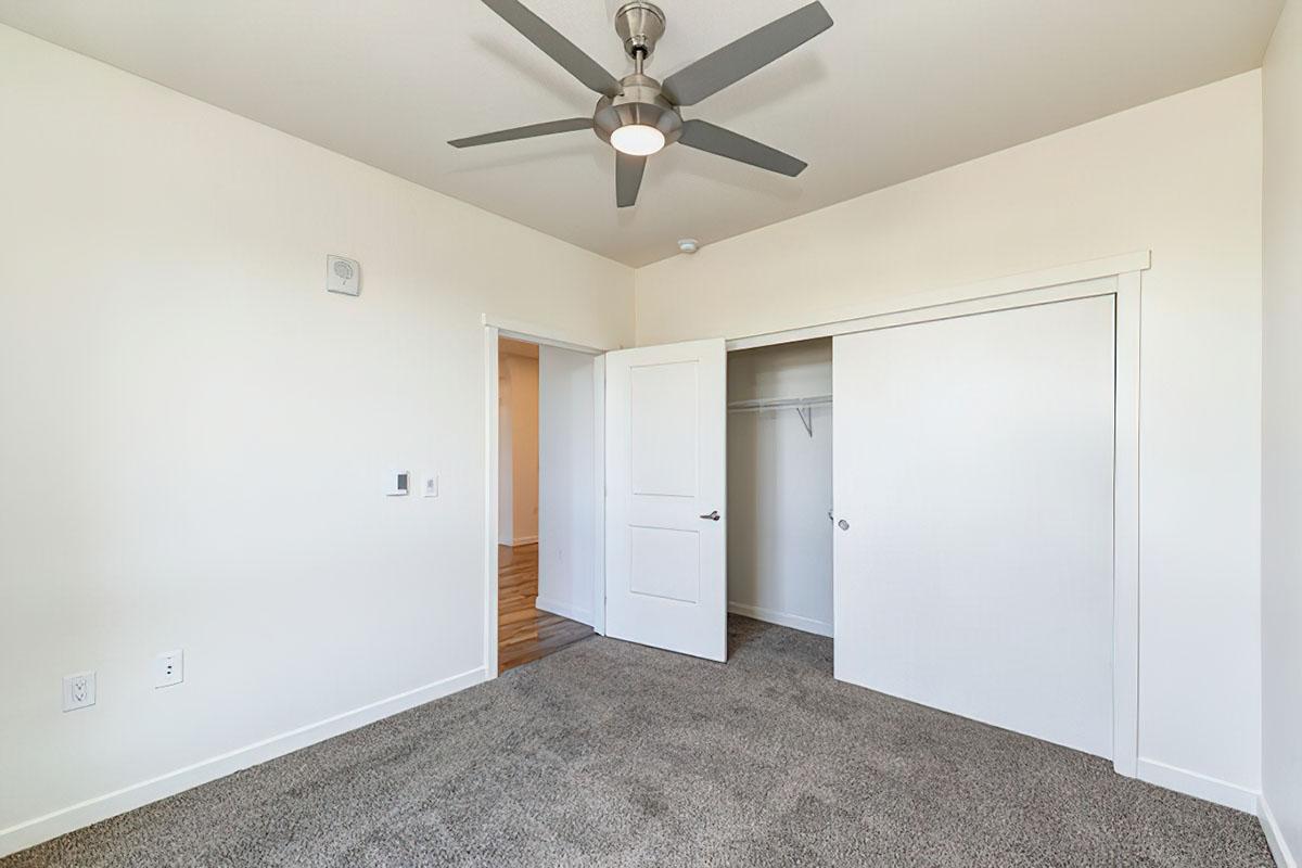A spacious, well-lit room featuring beige carpet flooring, a ceiling fan, and a neutral white wall.