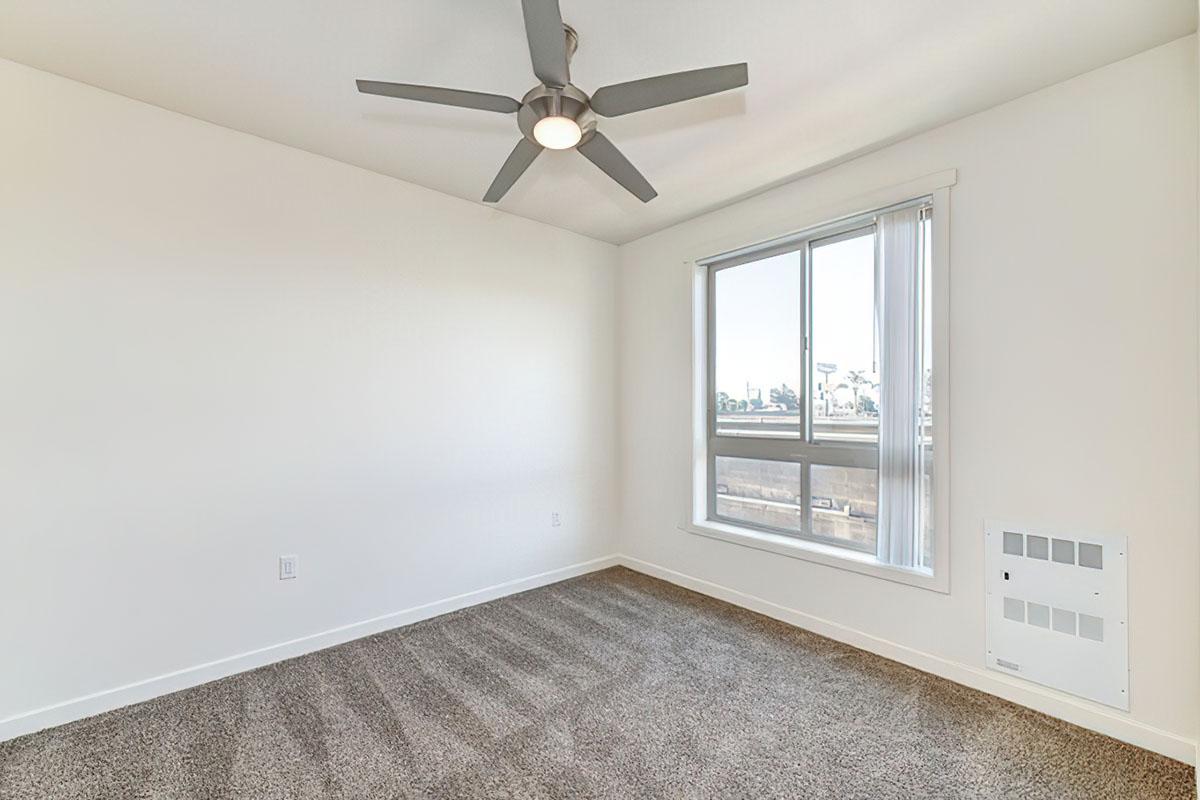 A brightly lit, empty room featuring a ceiling fan, a large window with vertical blinds, and beige carpet flooring