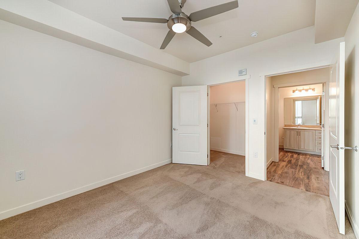 A spacious, empty room featuring a ceiling fan and light-colored walls. An open doorway leads to a bathroom area with modern fixtures.