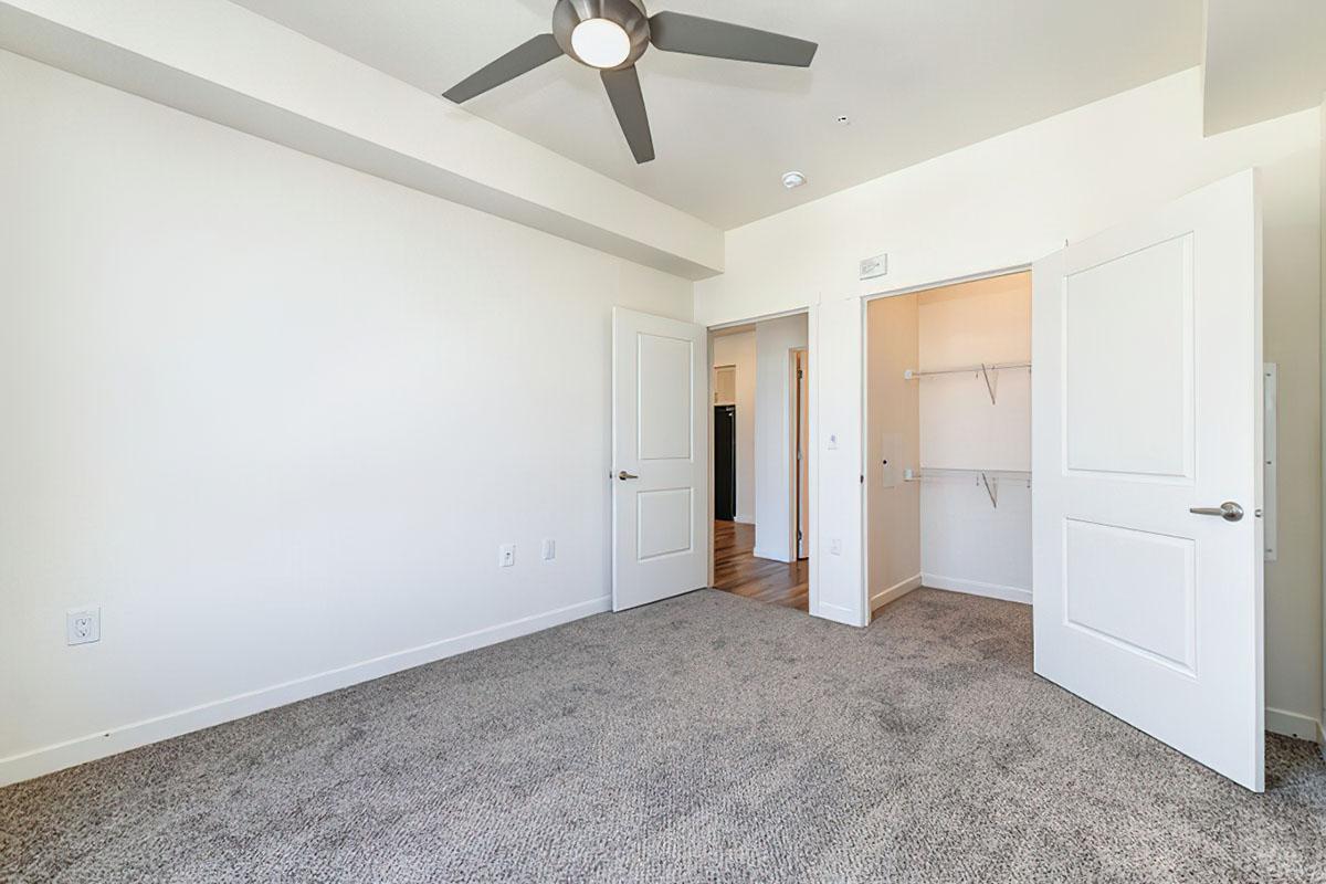 A spacious room with light-colored walls and carpet flooring. There is a ceiling fan and an open doorway leading to another area.