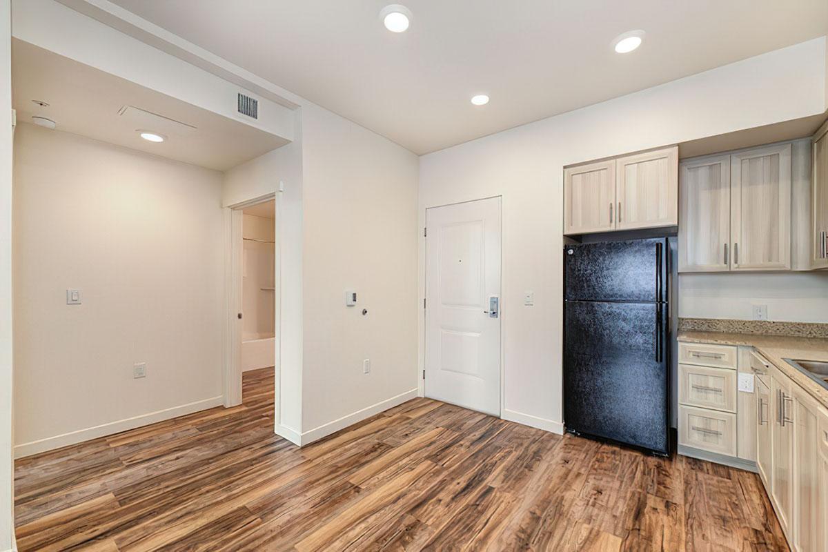 Contemporary kitchen and entryway of an apartment, featuring light-colored cabinets, a black refrigerator, and wood-style flooring.