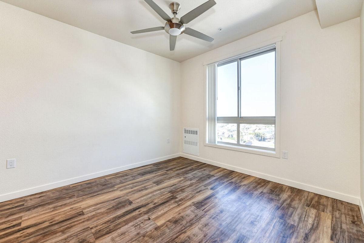 A vacant room with light-colored walls and a wooden floor. A ceiling fan is mounted in the center.