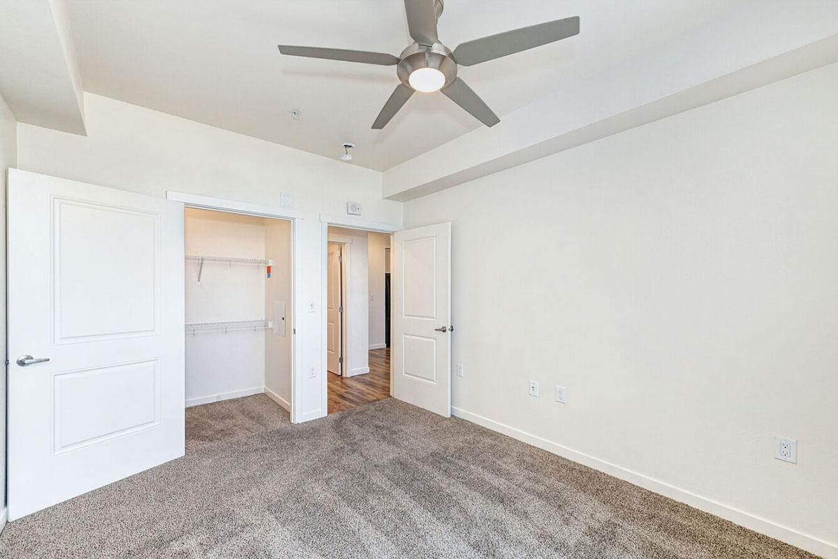 A well-lit, empty bedroom featuring soft beige walls and a plush carpet. The room has two open white doors leading to a closet and another space.