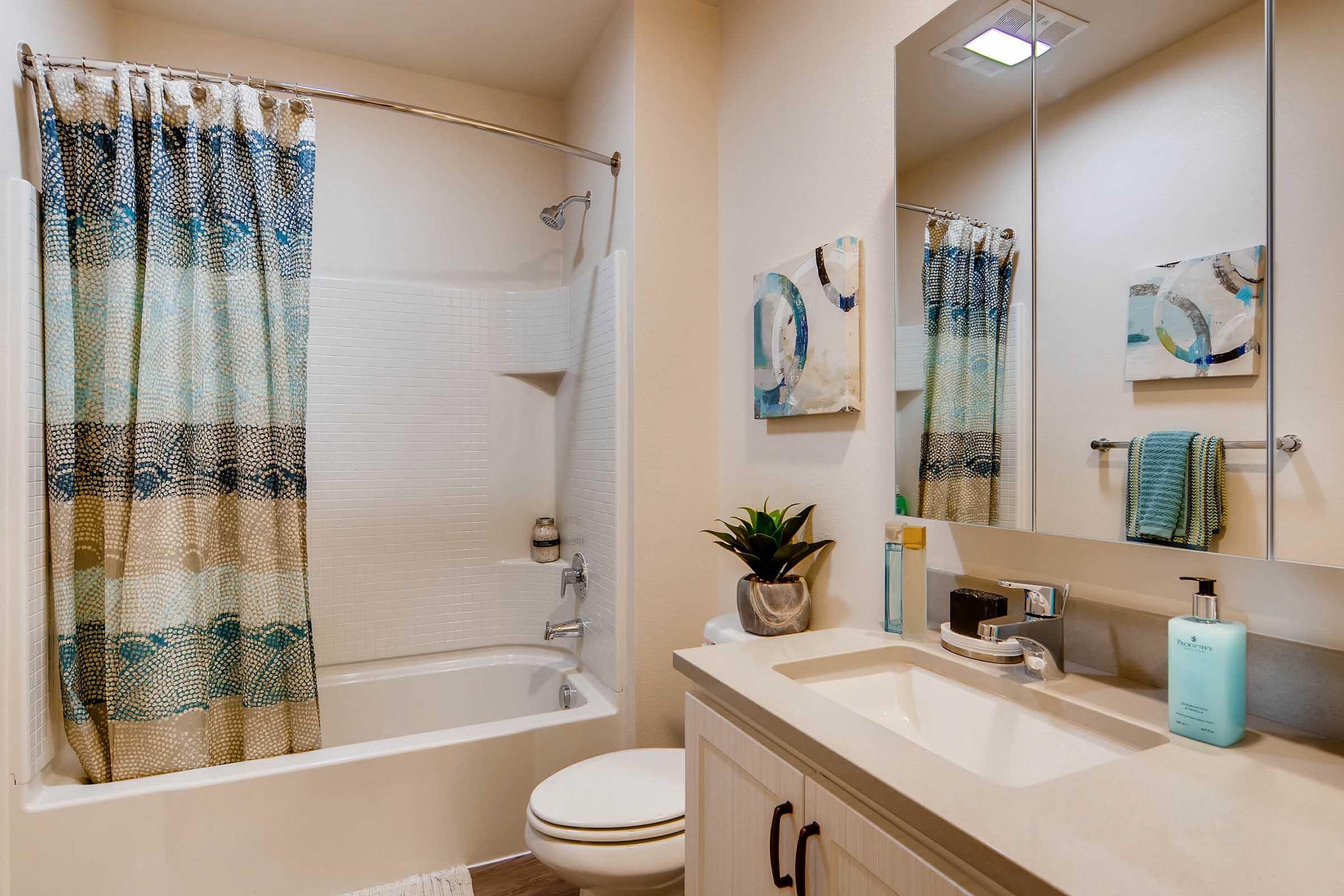 A well-lit bathroom featuring a white bathtub with a shower, a decorative shower curtain with blue and beige patterns, a light-colored vanity with a sink, a mirror above, and a small plant. Decorative art pieces are displayed on the wall, adding a modern touch to the space.