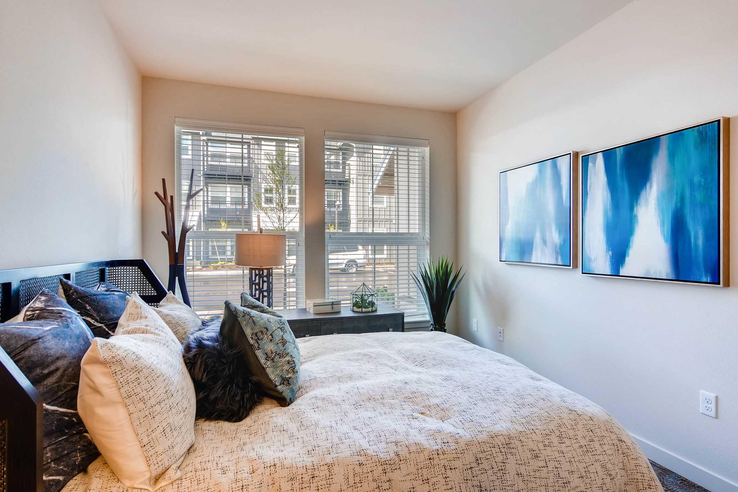 A bright and cozy bedroom featuring a comfortable bed with decorative pillows. There's a nightstand with a lamp next to the bed, and large windows letting in natural light. Two abstract blue paintings adorn the wall, and a potted plant adds a touch of greenery to the space.