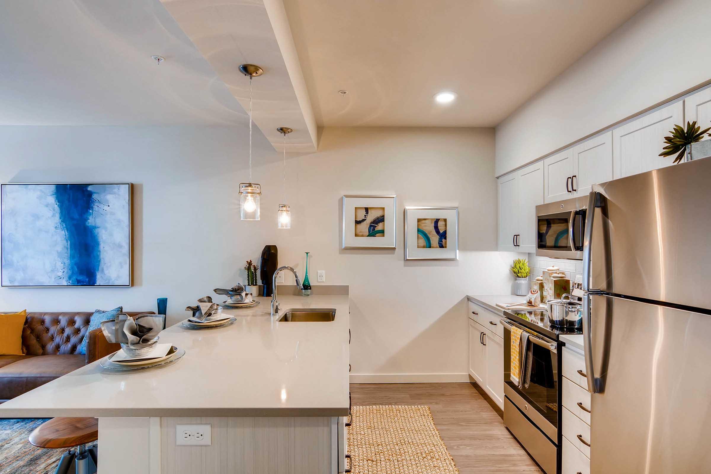 Modern kitchen and living area featuring a sleek design. The kitchen has a white countertop, stainless steel appliances, and pendant lighting. A cozy brown couch is visible in the background, along with abstract art on the walls and decorative items on the counter. Bright and inviting atmosphere.