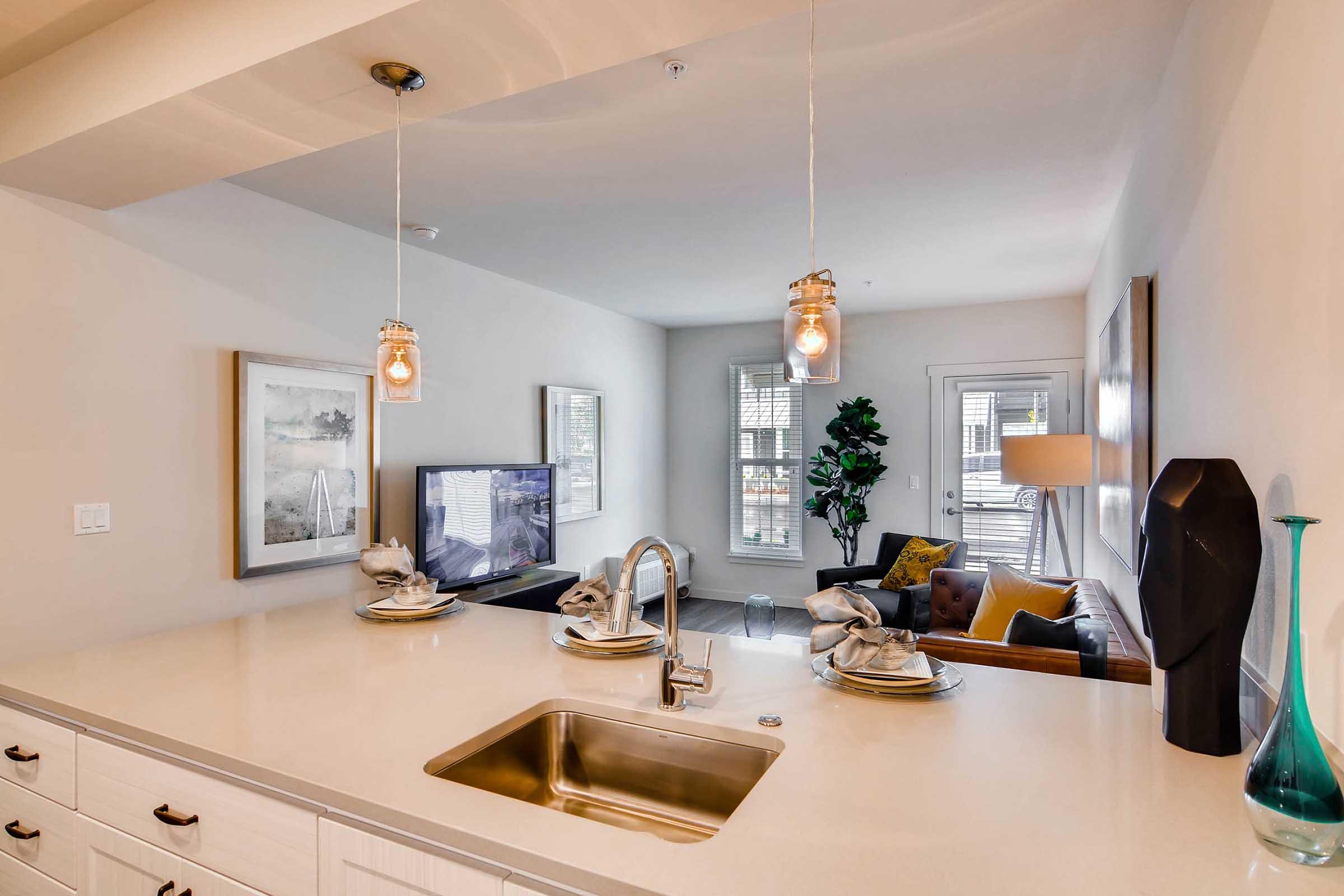 Modern kitchen and living room area featuring a sleek countertop with a sink and decorative place settings. A television is visible, alongside a sofa with cushions. There are large windows allowing natural light, a potted plant, and contemporary light fixtures hanging above the countertop, creating a warm and inviting atmosphere.