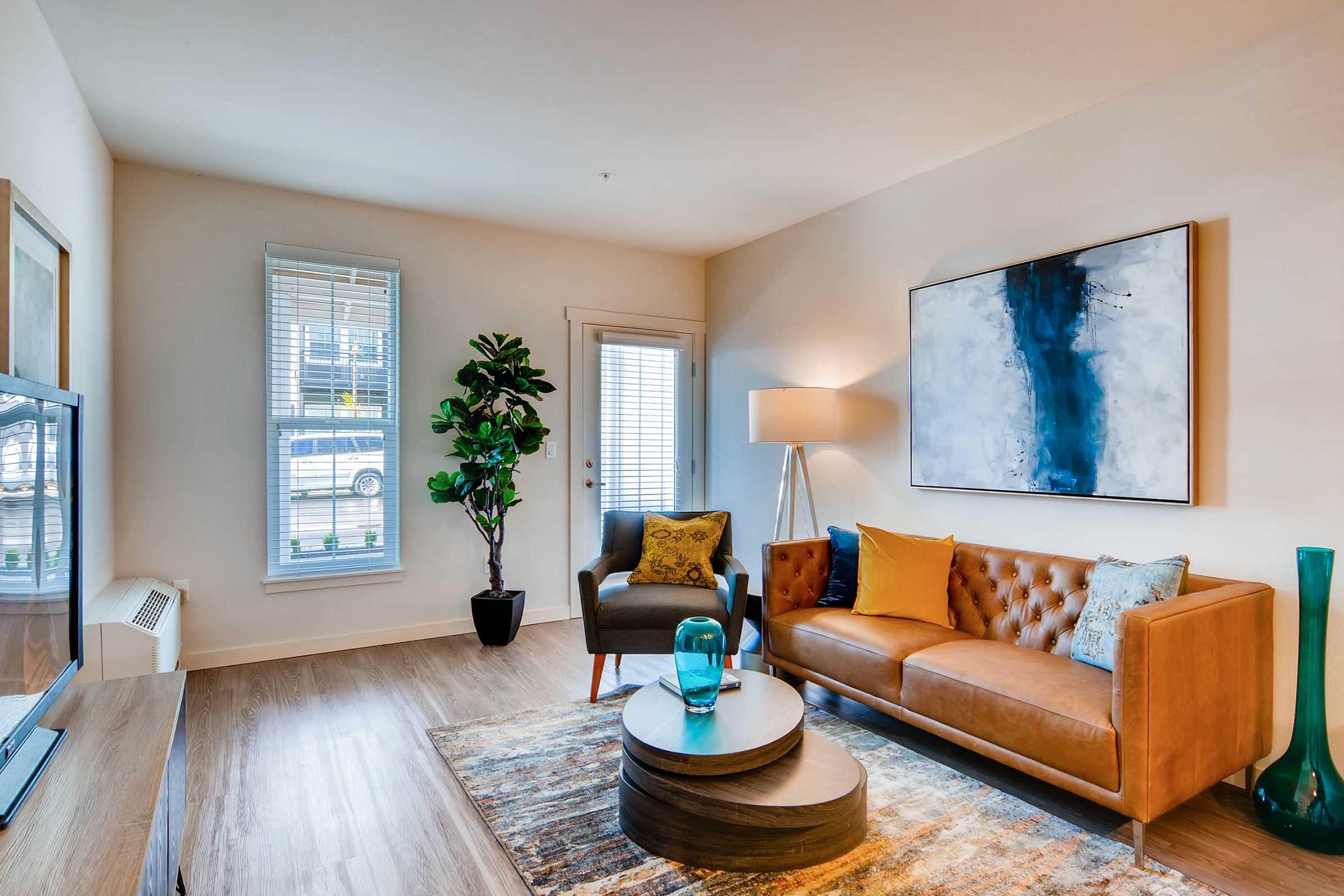 A cozy living room featuring a brown leather sofa with colorful cushions, a modern armchair, a round coffee table, and decorative accents. Large windows allow natural light, and a tall plant adds a touch of greenery. The walls are painted in soft, neutral tones, complemented by a contemporary piece of artwork.