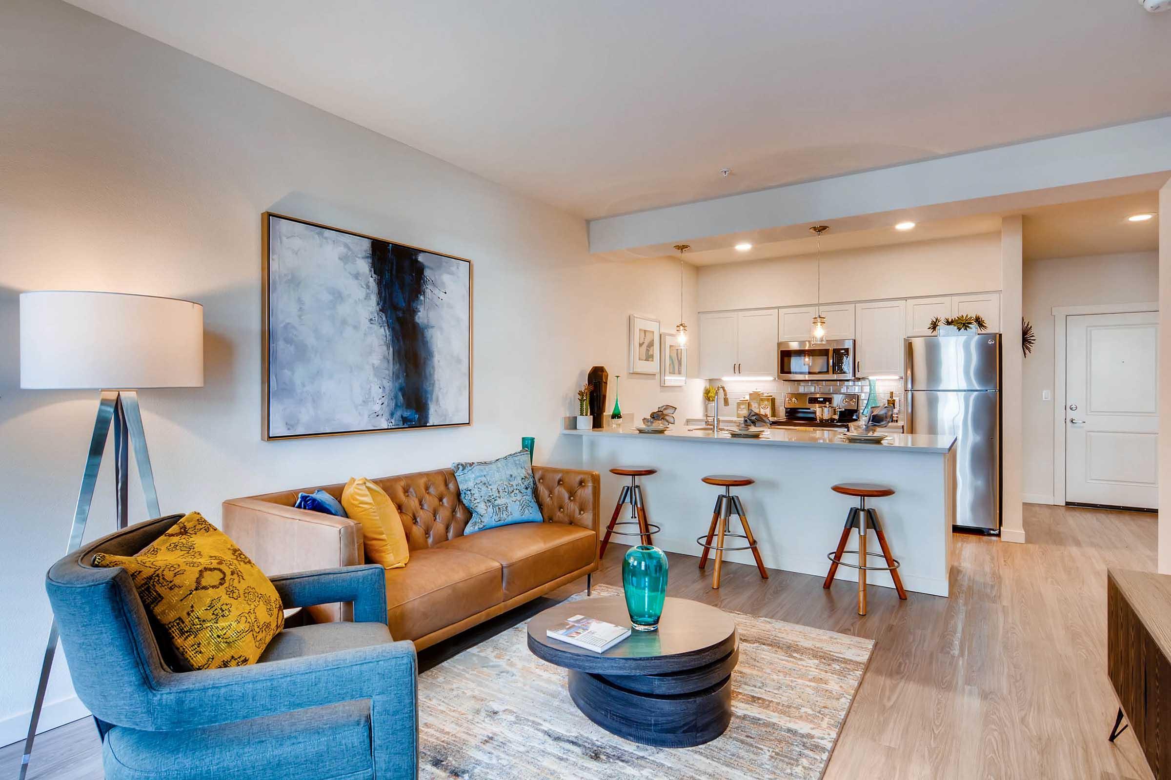 A modern living room featuring a brown tufted sofa with colorful pillows, a blue armchair, and a round coffee table with a green vase. In the background, there's a kitchen with stainless steel appliances, bar stools, and a light color scheme. The space is well-lit and stylishly decorated.