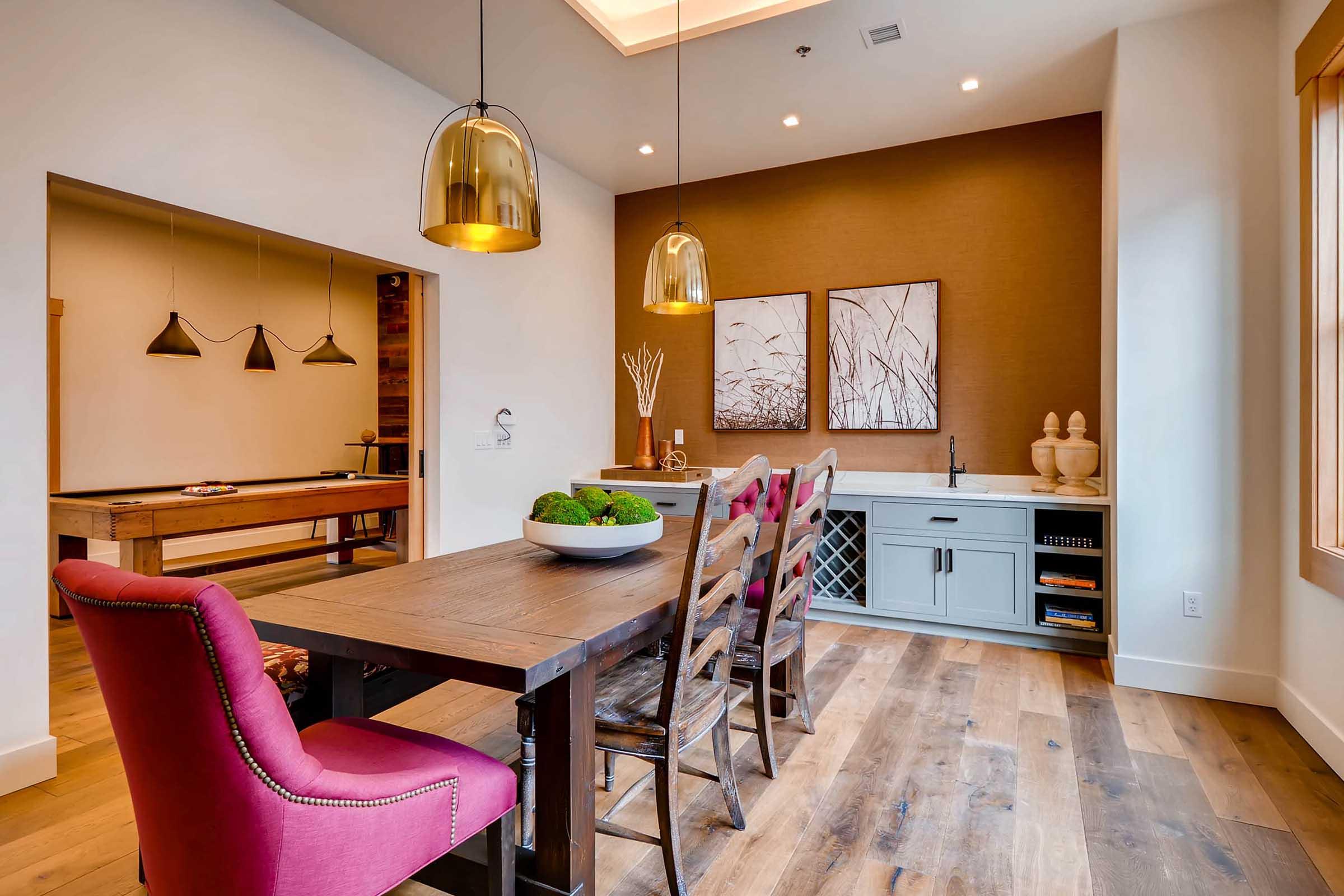 A modern dining area featuring a spacious wooden table surrounded by high-backed chairs. Soft lighting from pendant lamps above creates a warm atmosphere.