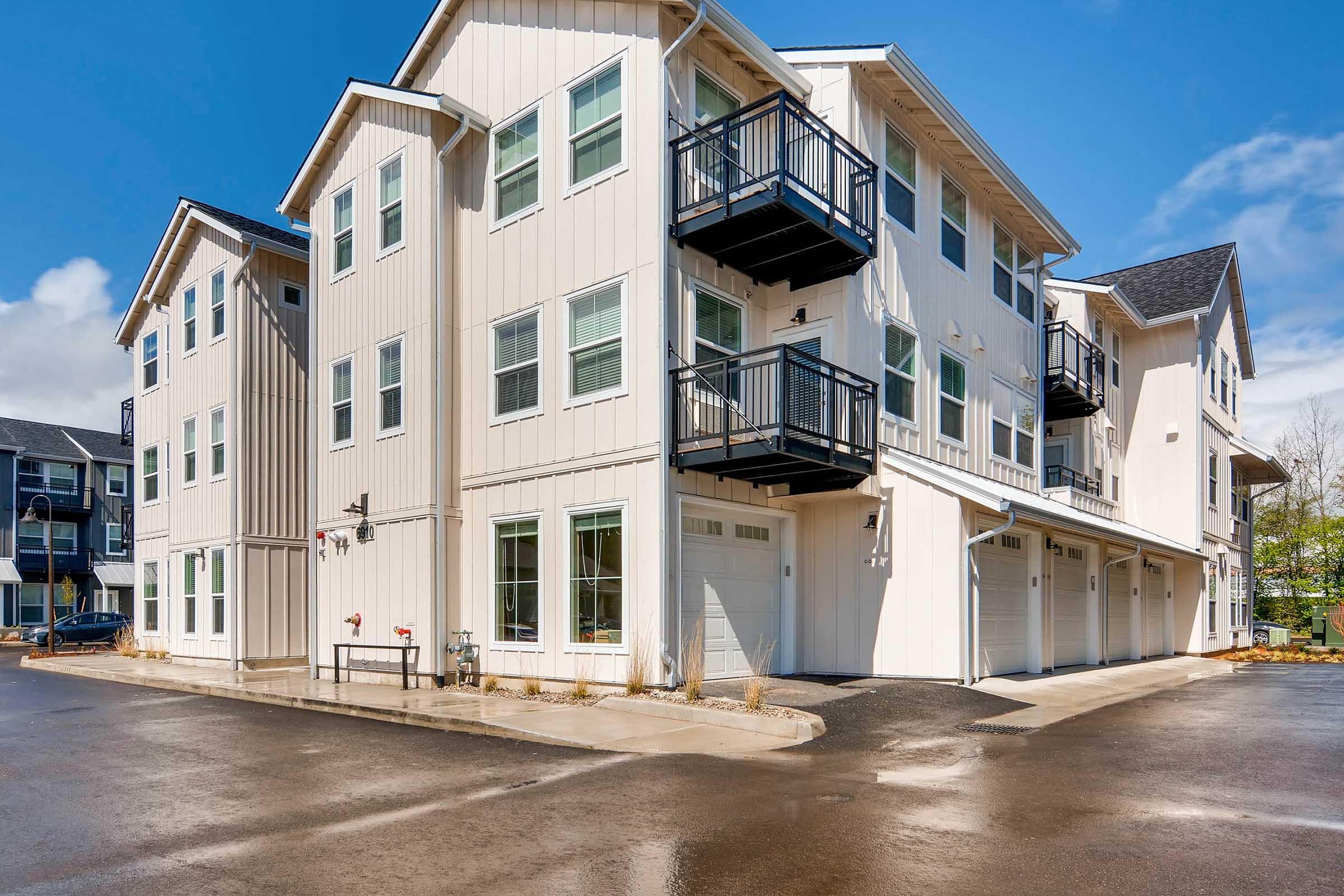 A modern three-story residential building featuring a light-colored exterior, multiple balconies, and garages at the ground level.