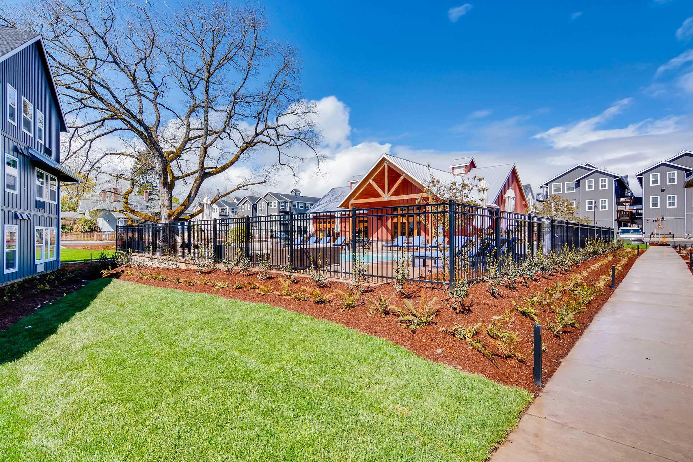 A landscaped area featuring a green lawn, a large fenced patio with a wooden structure, and several residential buildings in the background.