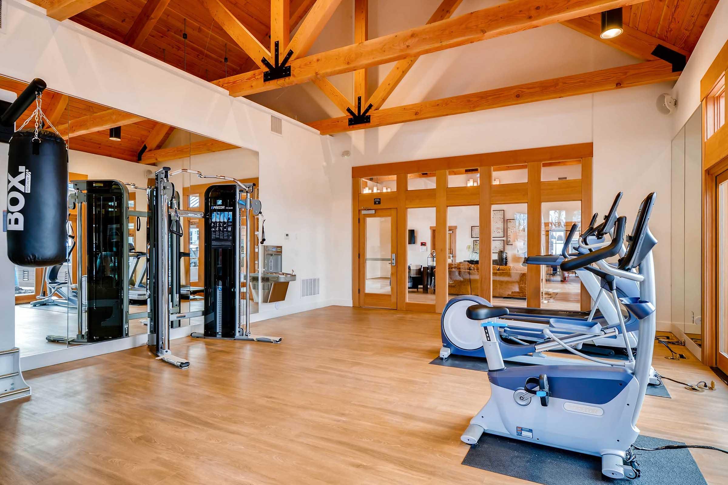 A spacious gym interior featuring large wooden beams, exercise equipment including a stationary bike and an elliptical machine, a boxing bag, and mirrored walls.
