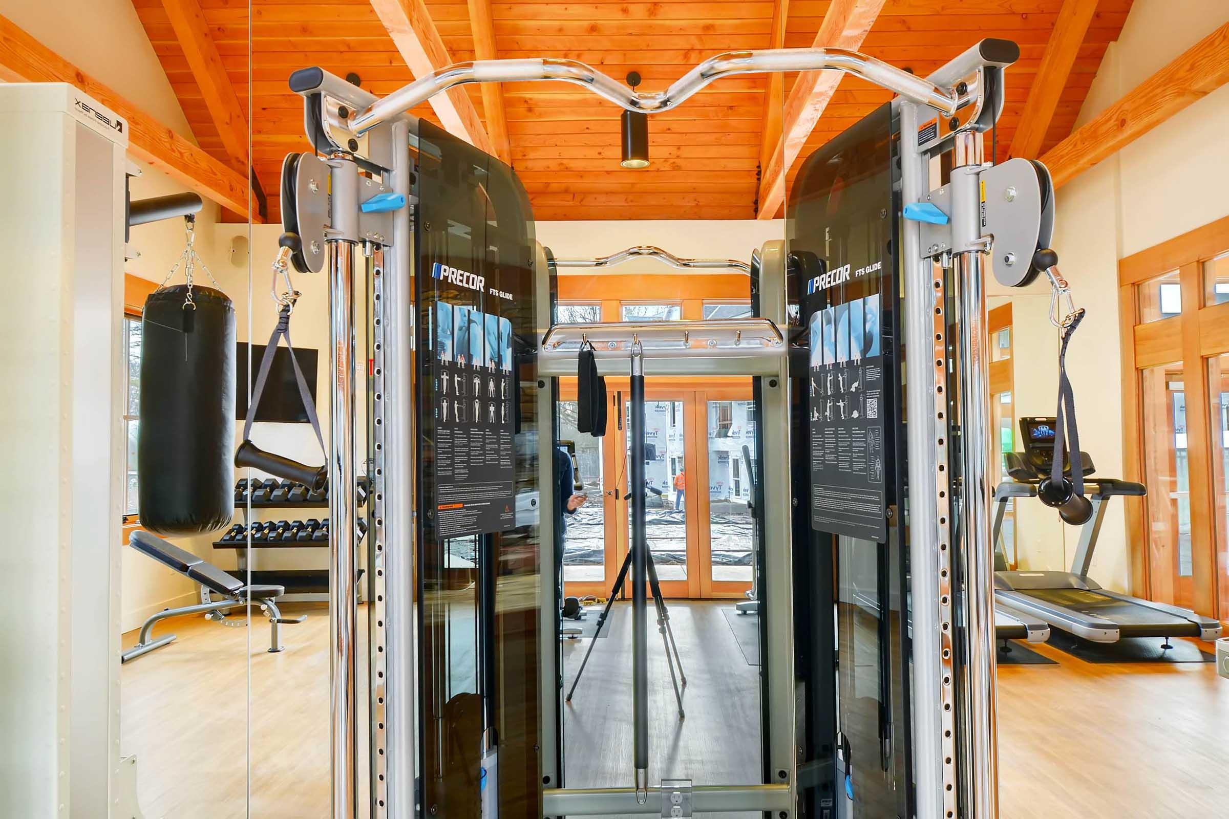 A modern gym interior featuring a cable machine in the center, a punching bag on the left, and various weight training equipment in the background.
