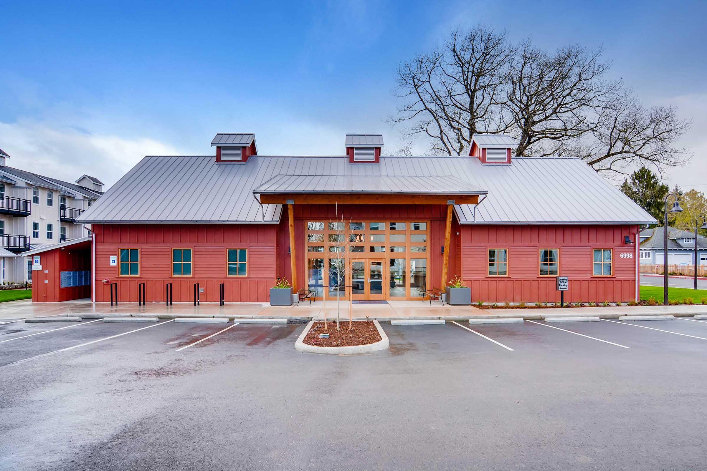 A modern, red barn-style building with a metal roof and large windows at the front. The structure features a central entrance with