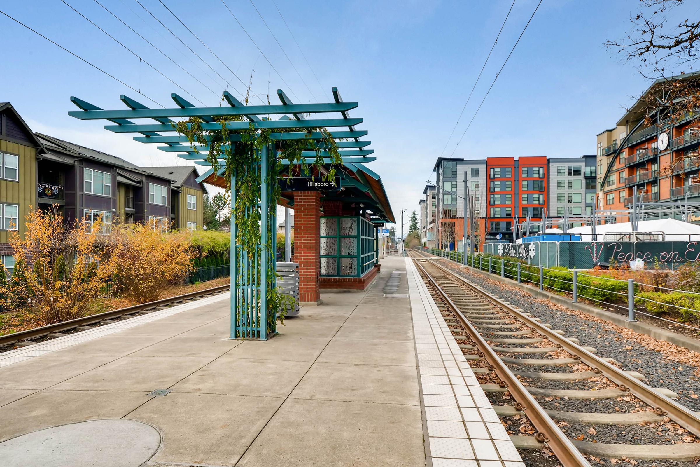 Railway station platform with a blue pergola, surrounded by greenery and residential buildings.