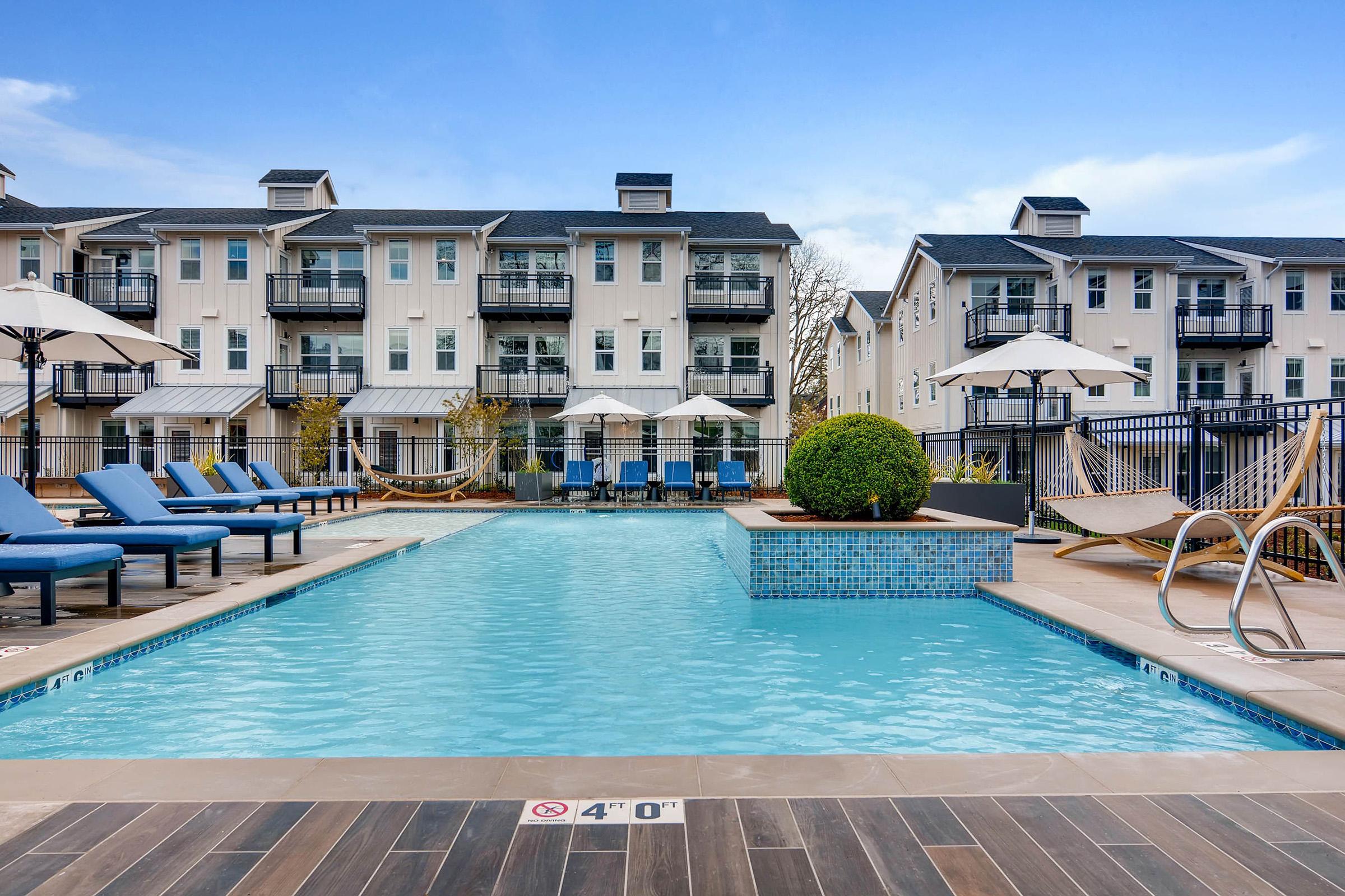 A modern outdoor swimming pool surrounded by lounge chairs and umbrellas, with a backdrop of stylish apartments featuring balconies.