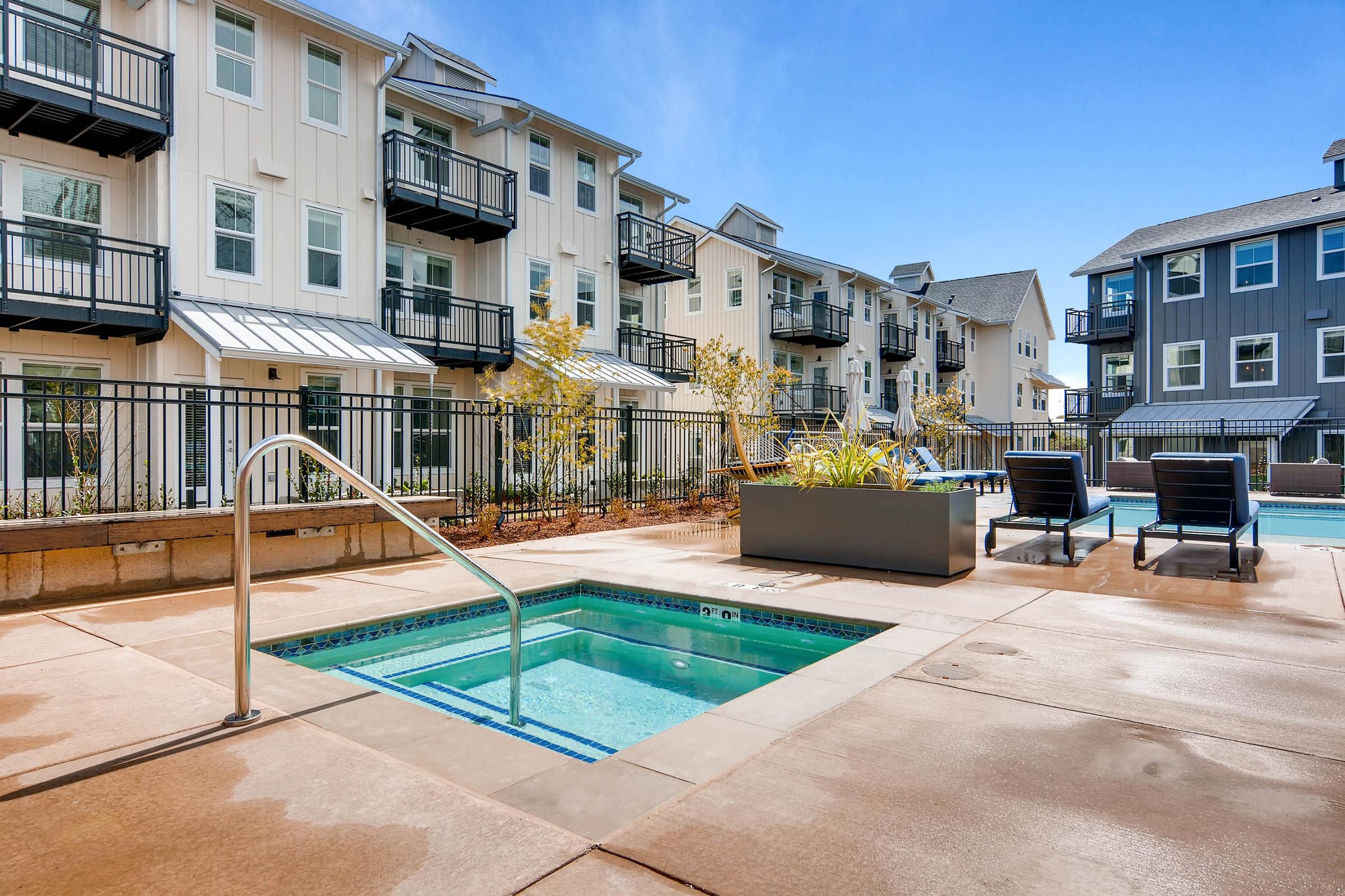 A sunny outdoor area featuring a hot tub and lounge chairs next to a swimming pool.