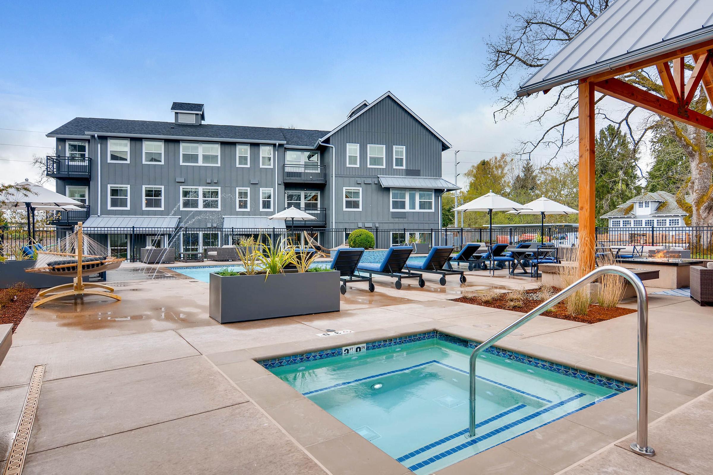 A well-maintained outdoor pool area featuring a hot tub, lounge chairs, and shaded umbrellas.