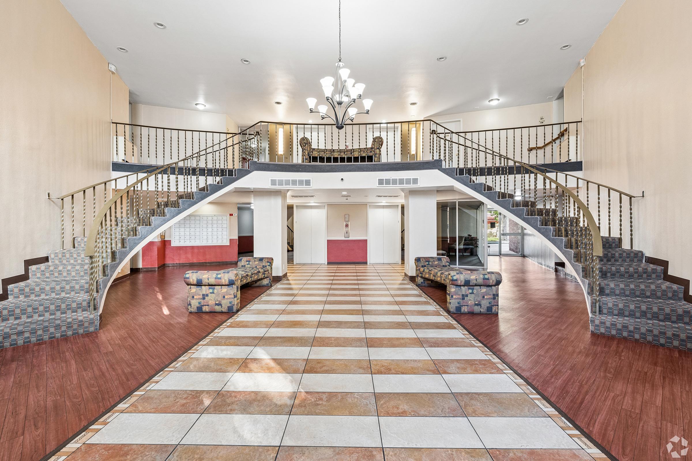 Interior view of a spacious lobby featuring a grand staircase with decorative railings. The floor has a checkered pattern with tiles and wood accents. There are two patterned sofas on either side of the entrance, and a chandelier hanging from the ceiling. The walls are painted in light colors, creating an inviting atmosphere.