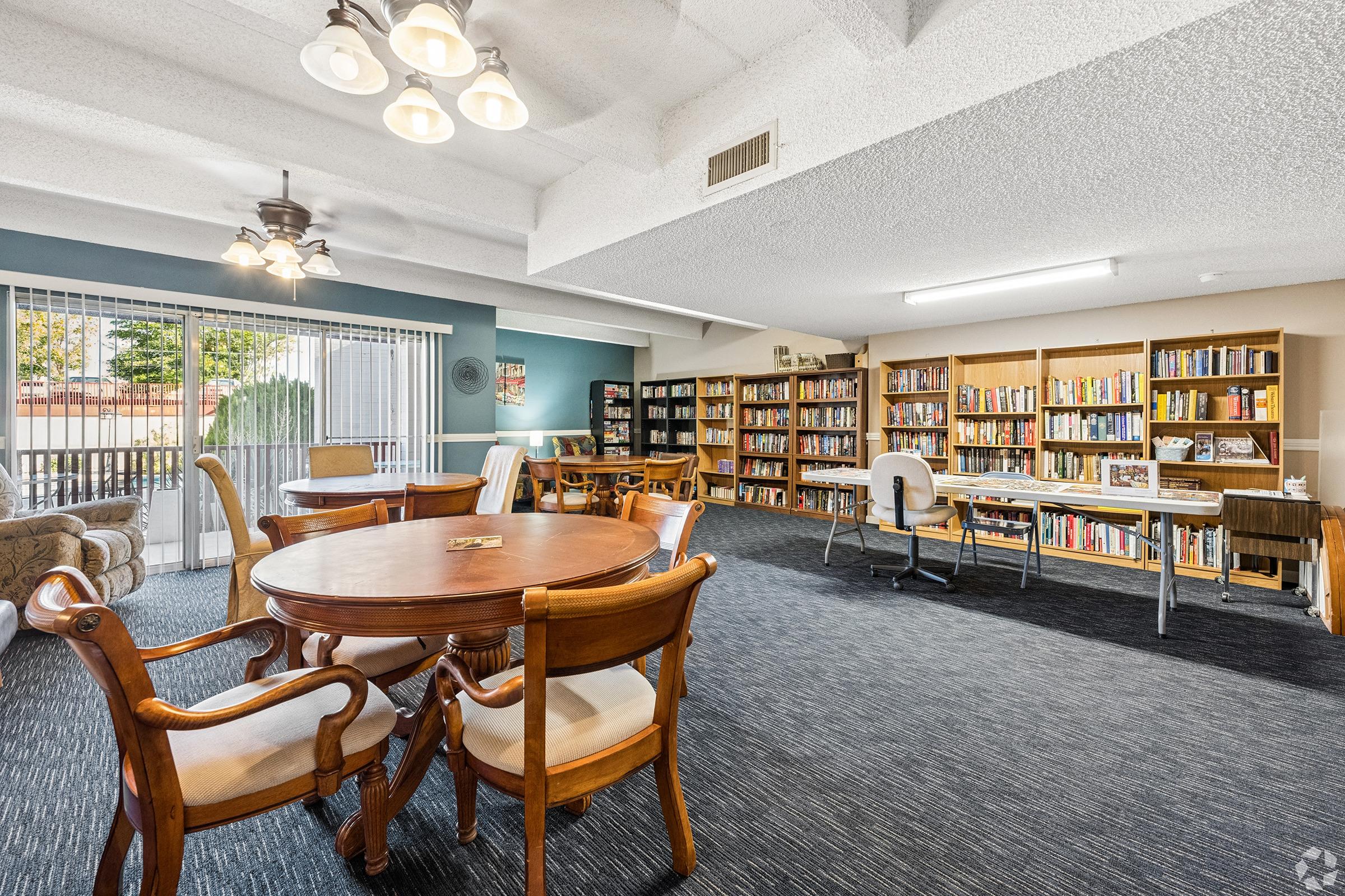 A cozy library or reading room featuring wooden tables and chairs, bookshelves lined with books, and large windows allowing natural light. The space has a warm atmosphere with a mix of comfortable seating, offering a welcoming environment for reading or studying.