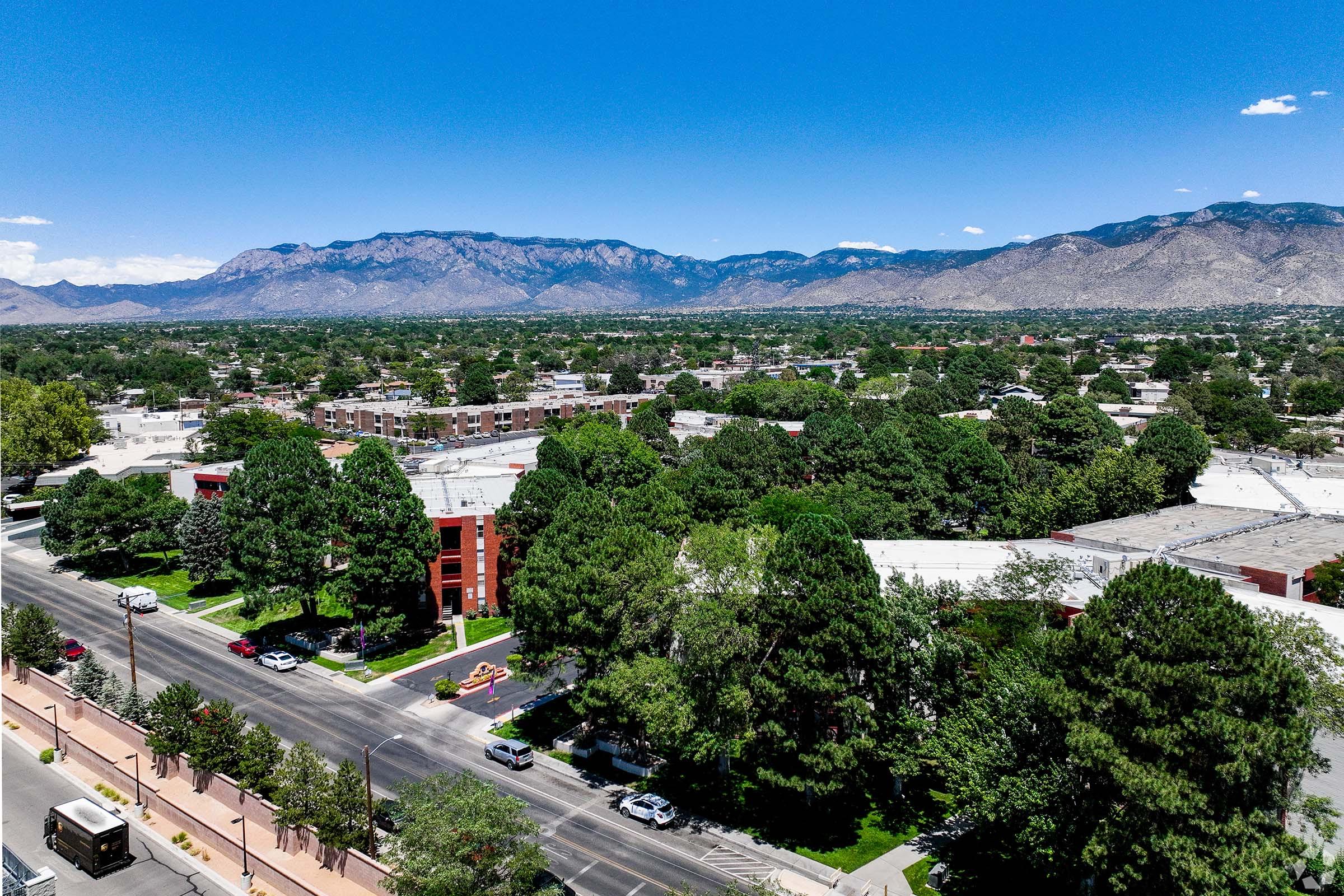 Aerial view of a cityscape featuring residential buildings, lush green trees, and a wide road lined with parked cars. In the background, majestic mountains rise against a clear blue sky, showcasing a scenic blend of urban and natural landscapes.