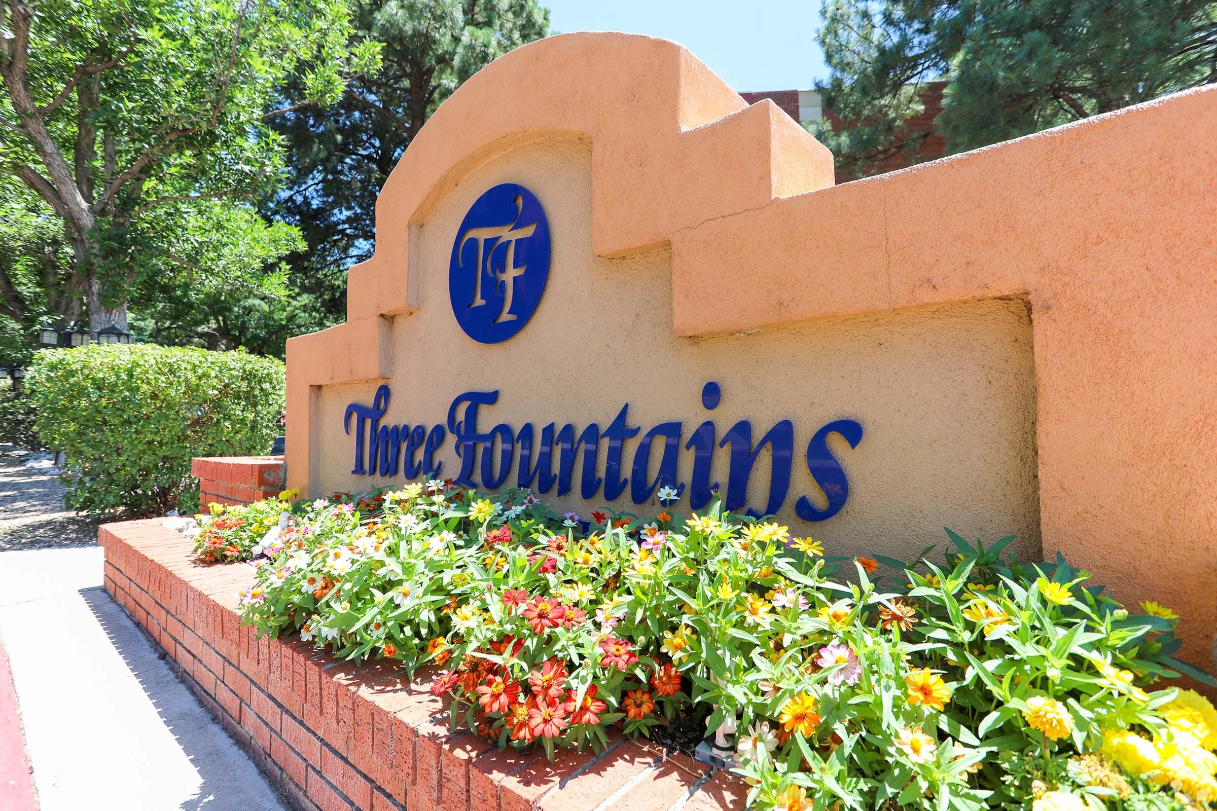 Sign for "Three Fountains" with a stylized logo, surrounded by colorful flower beds. The sign features a warm orange background with blue lettering, set against a backdrop of greenery.