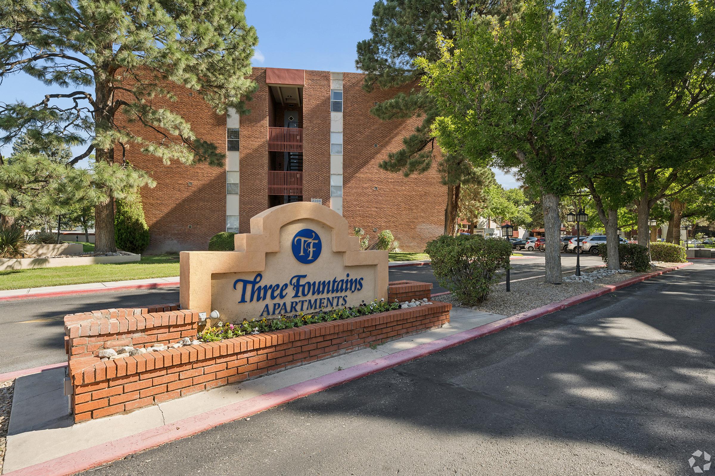 Sign for Three Fountains Apartments, featuring the name in large, prominent letters. The background includes a brick structure with landscaping, including trees and shrubs. The pathway is lined with red curbing, creating a welcoming entrance to the apartment complex.