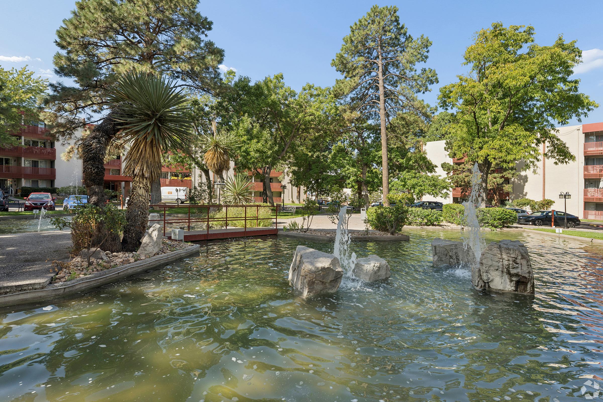 A tranquil courtyard scene featuring a small pond with water fountains and rocks, surrounded by lush green trees and shrubs. In the background, there are apartment buildings and parked cars, creating a peaceful residential atmosphere. Bright blue sky complements the greenery.
