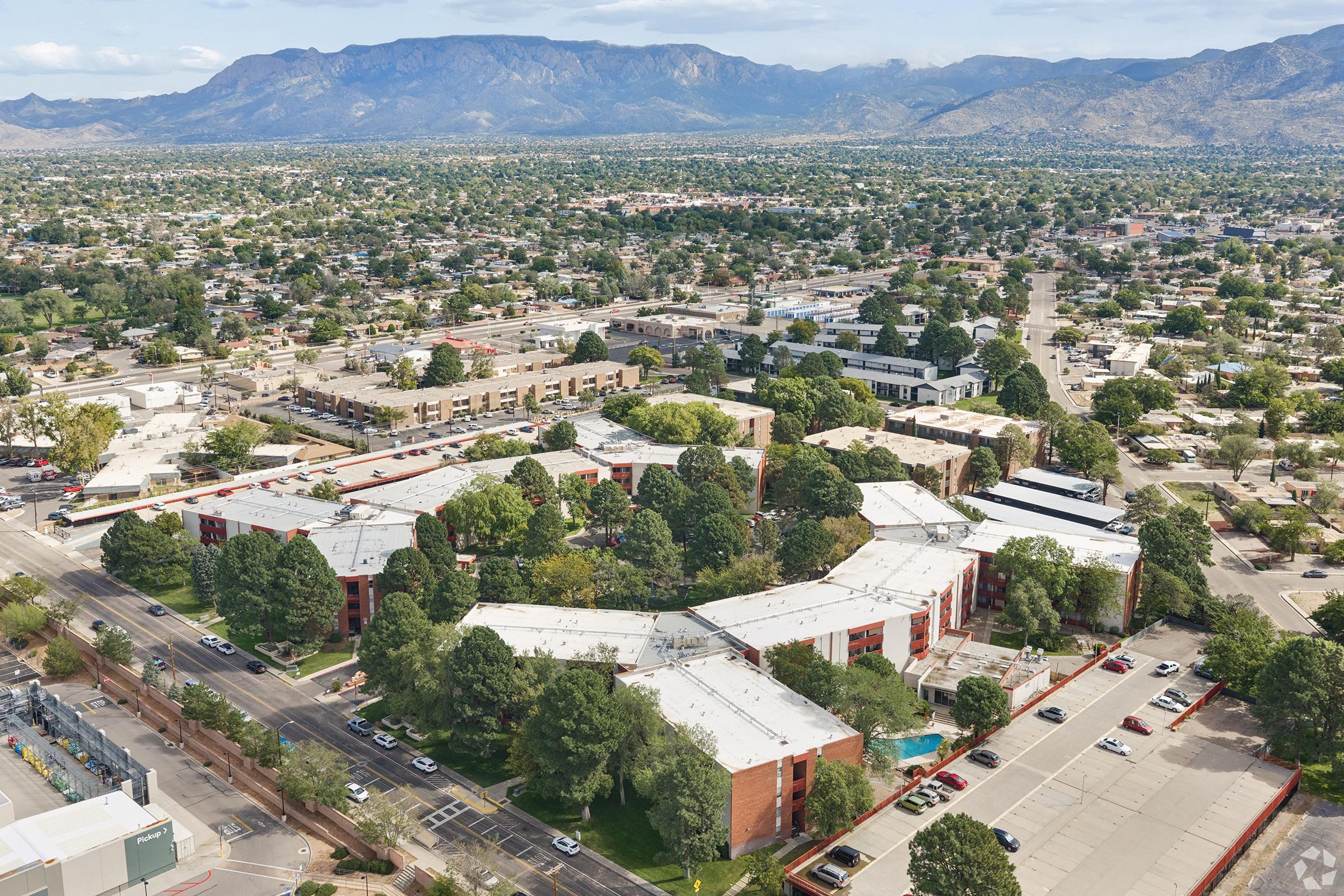 Aerial view of a suburban area featuring clusters of residential buildings, surrounded by trees and parking lots. In the background, mountains rise against a partly cloudy sky, while the foreground shows a layout of streets and vehicles. The overall scene captures a blend of urban and natural landscapes.