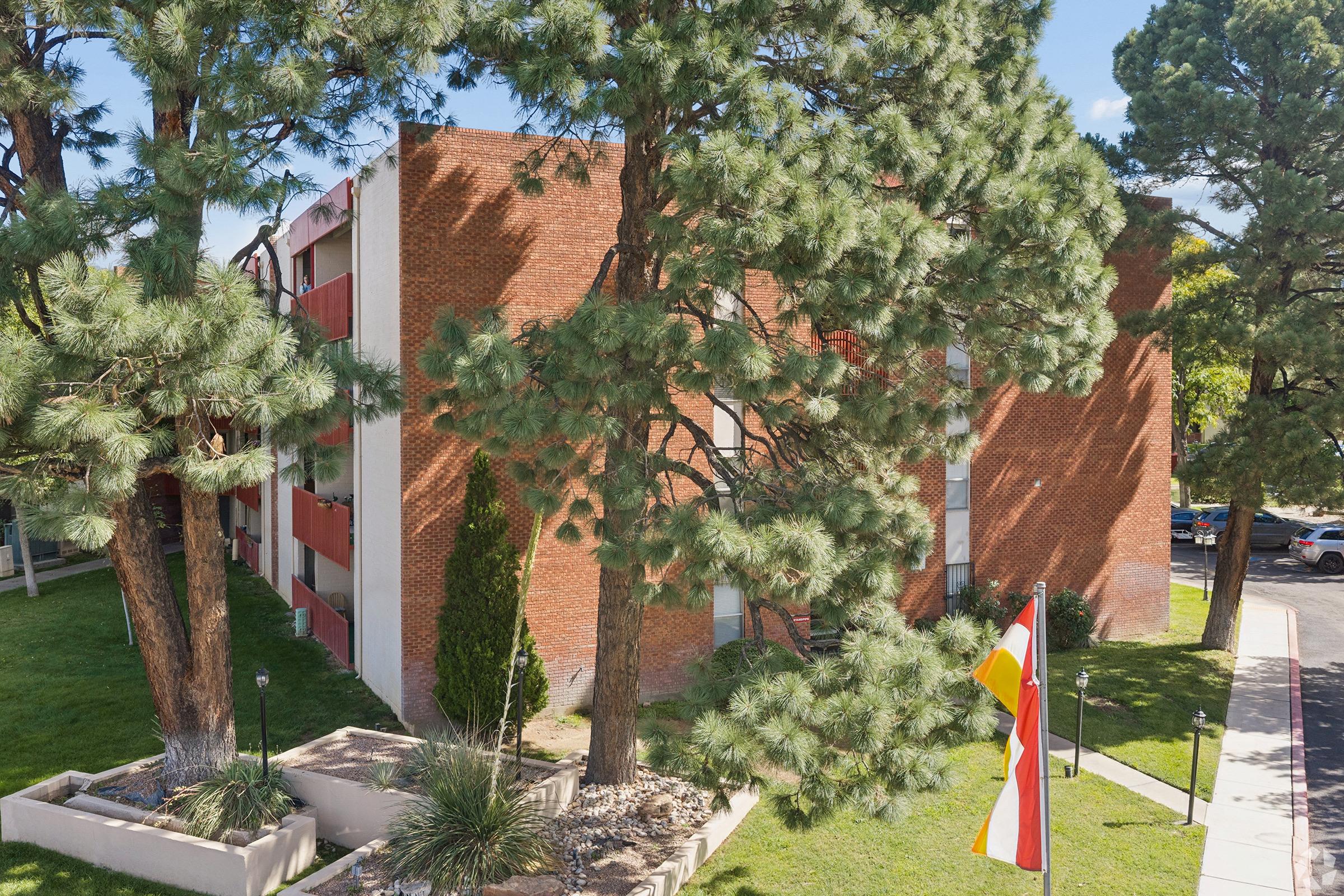 A view of a brick building partially obscured by tall pine trees. The building features multiple windows and balconies, with a well-maintained lawn in the foreground. In the foreground, a flagpole displays a flag. The scene is set on a sunny day, showcasing a clear blue sky and green foliage.