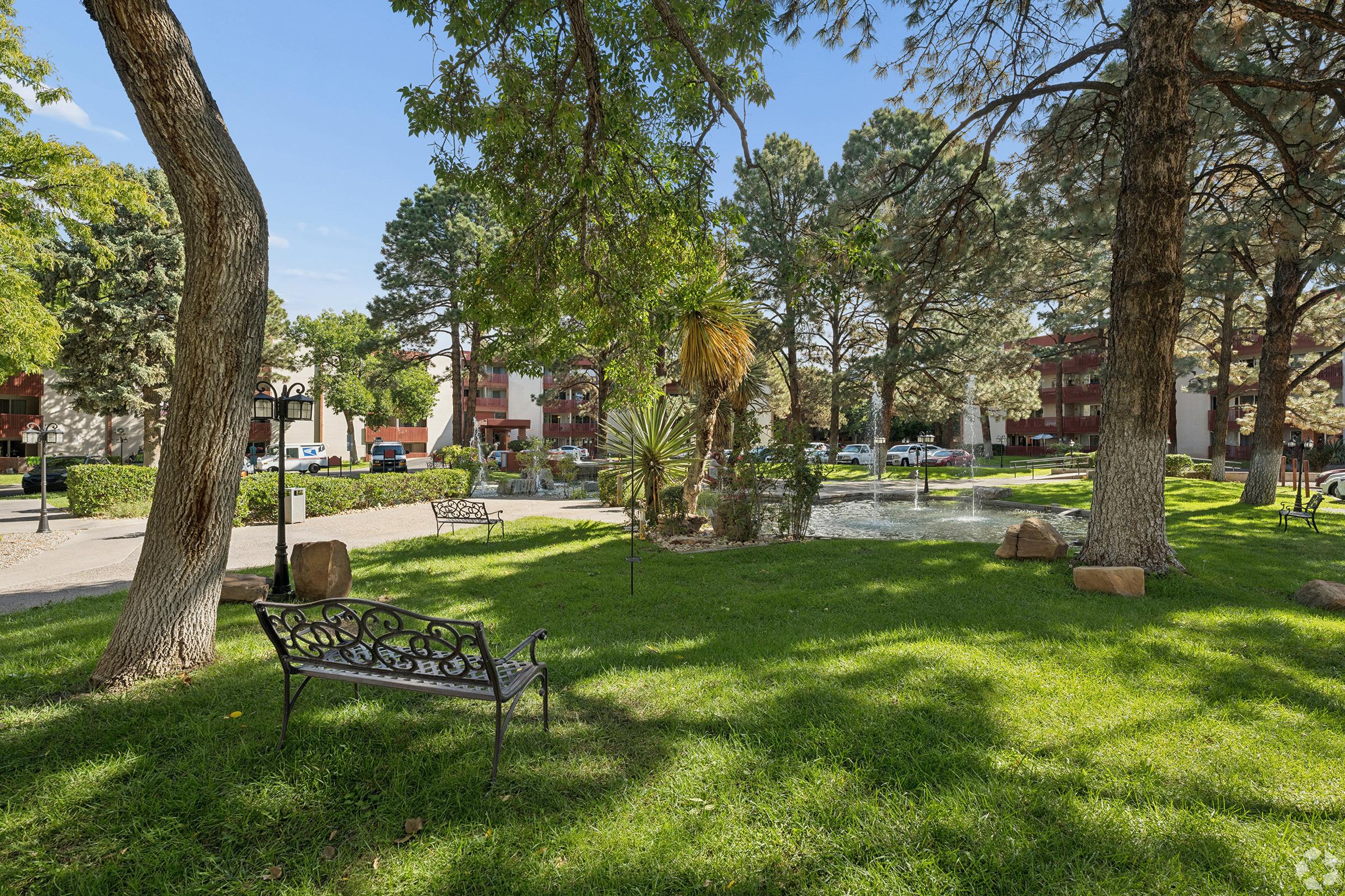 A peaceful outdoor park scene featuring green grass, tall trees, and a small pond. There are several benches scattered throughout the area, surrounded by well-maintained landscaping. In the background, residential buildings can be seen partially obscured by the trees, creating a serene atmosphere.