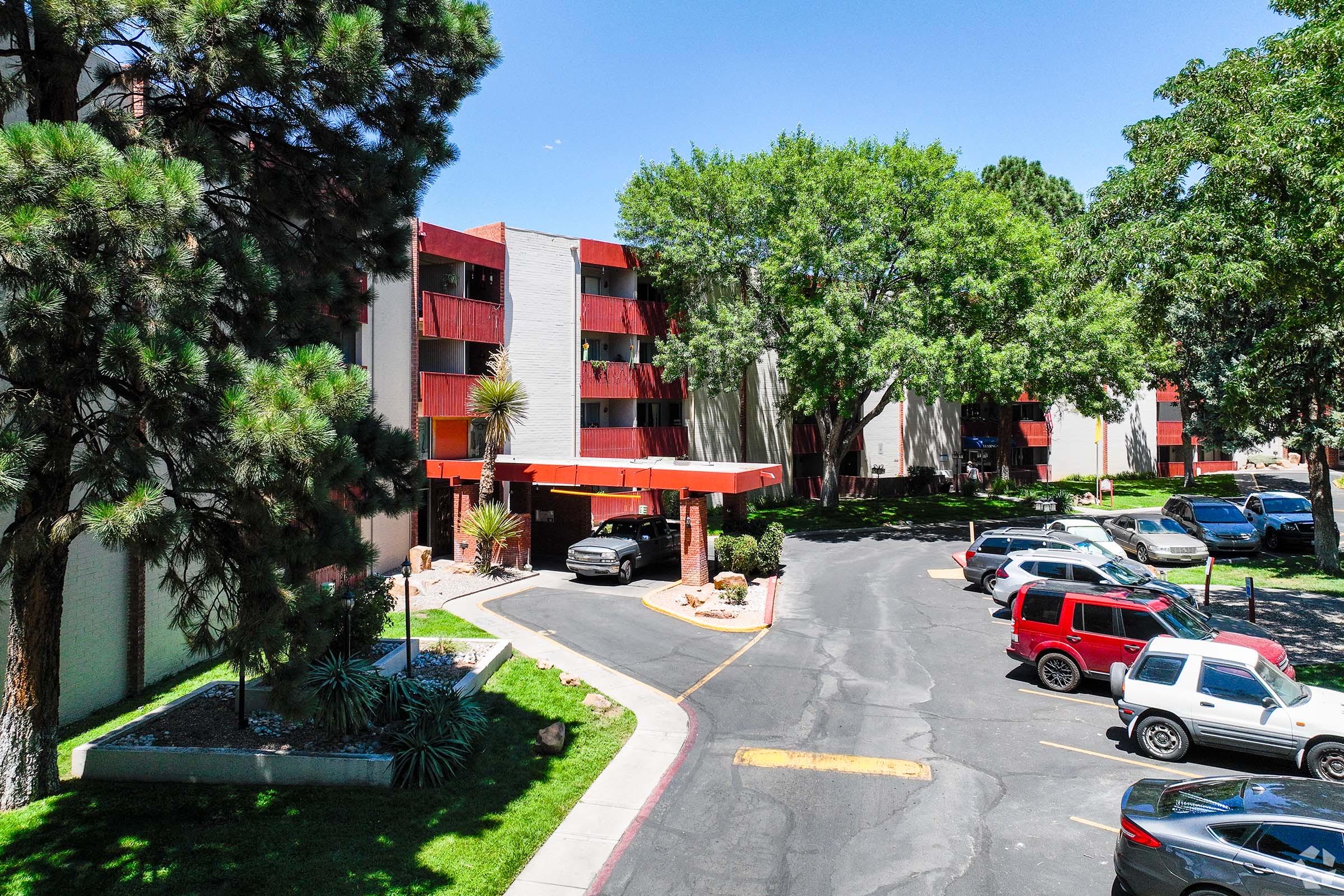 A view of a multi-story apartment complex featuring red and gray exteriors, surrounded by lush green trees. There are parking spaces filled with various cars along a winding driveway, leading to a covered entrance at the front of the building on a sunny day.