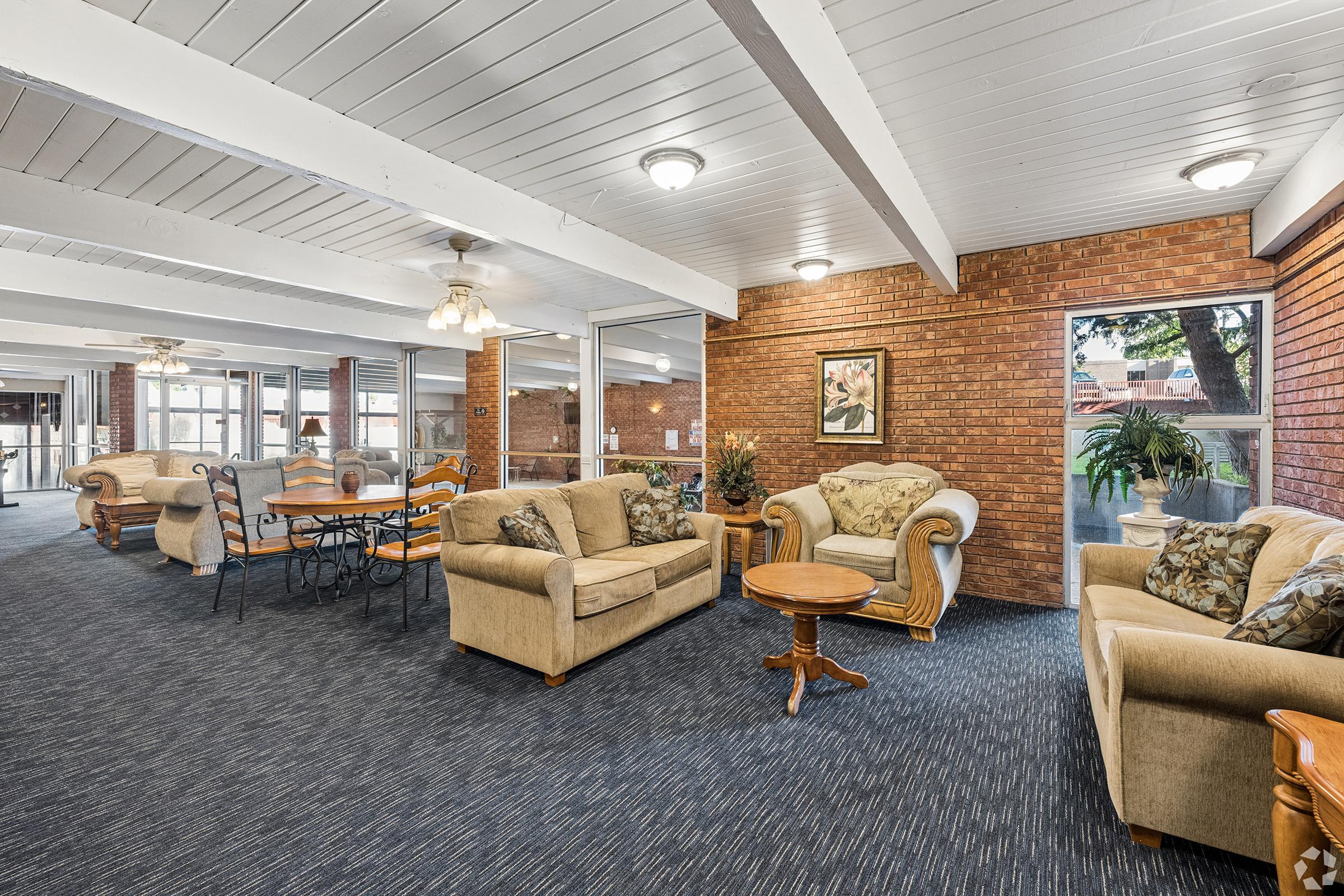 A cozy lounge area featuring beige sofas and a wooden coffee table. The space has wooden beams on the ceiling, a blue carpet, and brick walls. There's a dining area with a table and chairs in the background. Large windows allow natural light to brighten the room, with plants adding a touch of greenery.