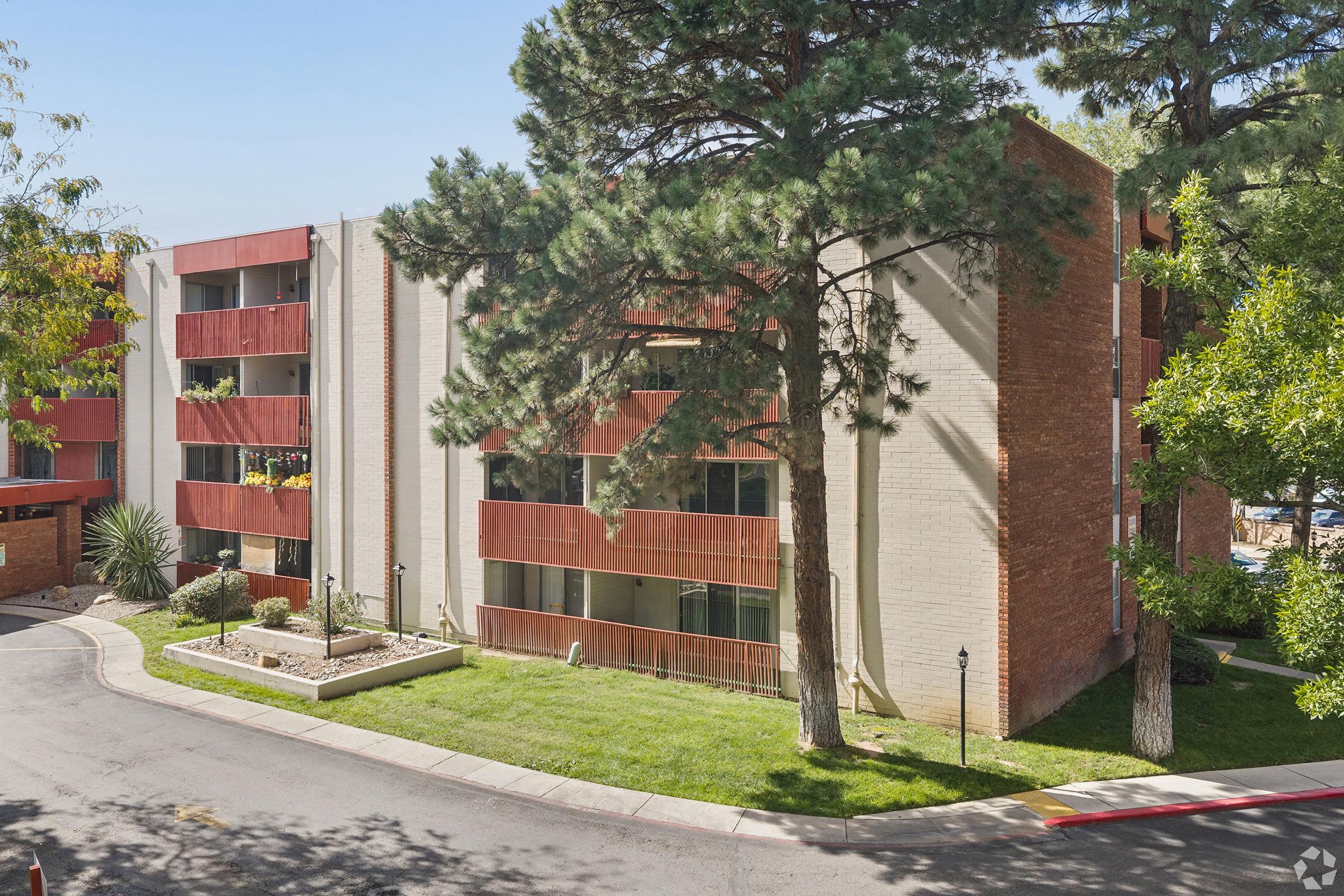 A multi-story apartment building with a mix of red and beige exteriors. Surrounded by greenery, including trees and shrubs. The area features a curved driveway leading to the entrance, with a well-maintained lawn in front. Bright, sunny day with a clear blue sky.