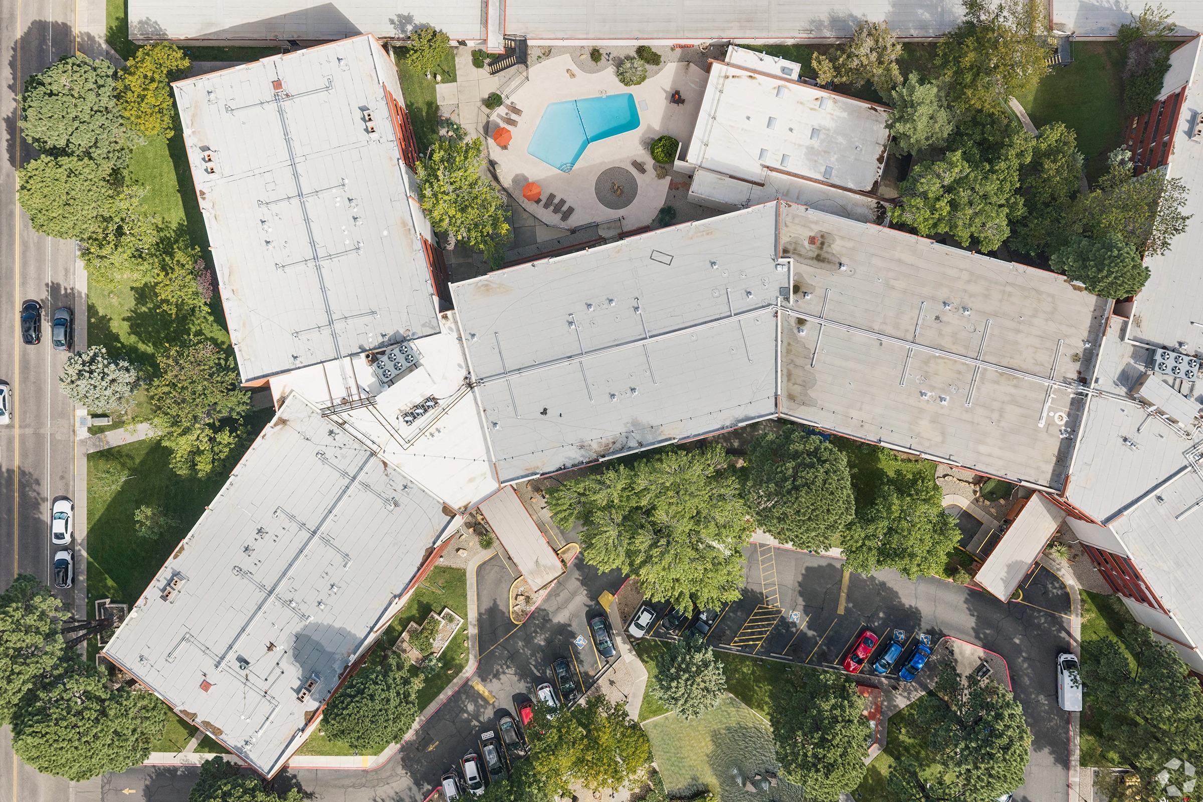 Aerial view of a complex featuring multiple buildings arranged in an interconnected layout. In the center, a small swimming pool is surrounded by a patio area with seating. Lush greenery and trees are visible around the buildings, while a parking lot with several cars is situated nearby.