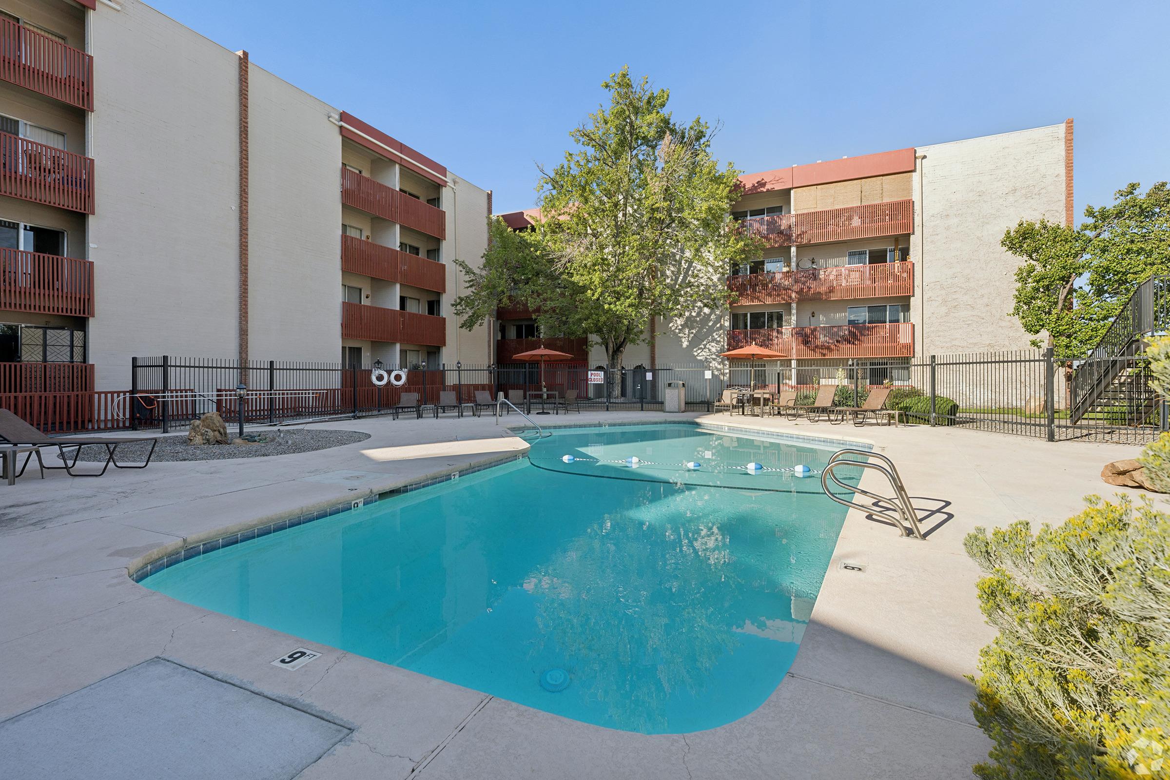 A clear blue swimming pool surrounded by landscaped areas, with lounge chairs and poolside umbrellas. The pool is situated in a courtyard of a multi-story apartment building with balconies. The scene is set on a sunny day, featuring a few trees providing shade.