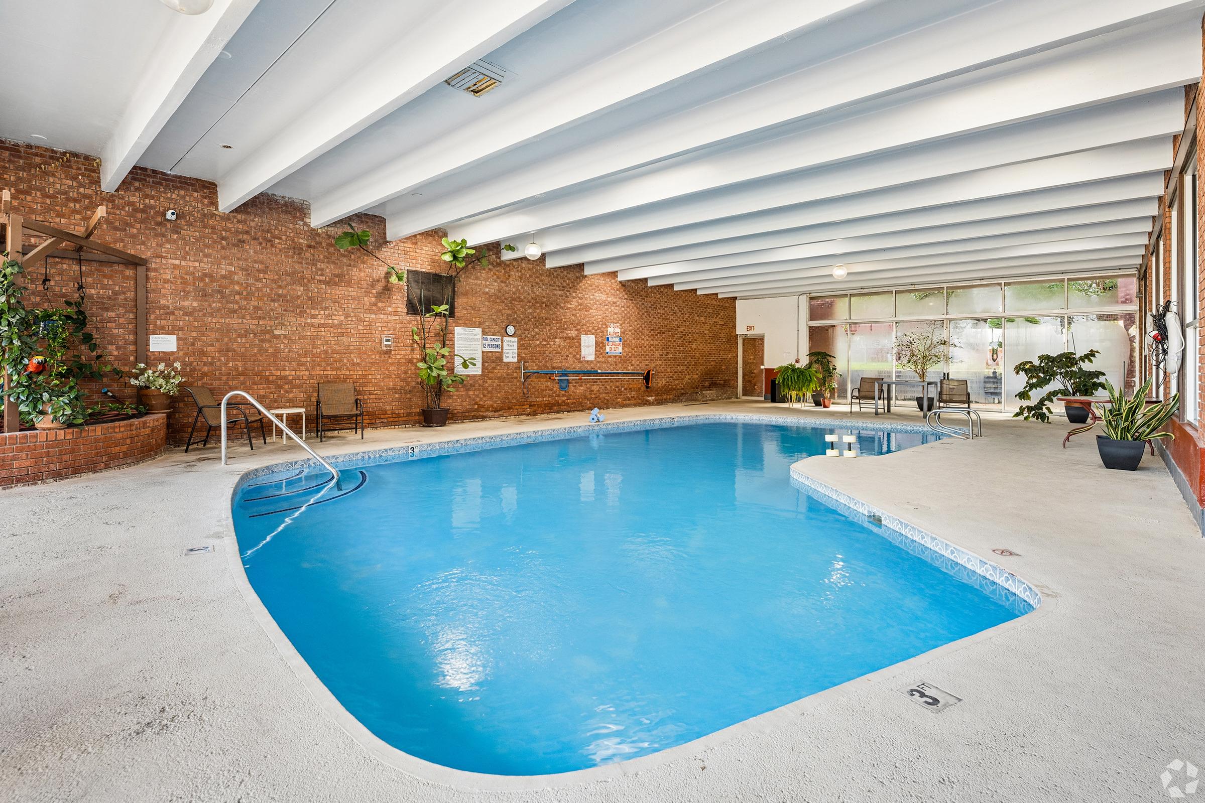 Indoor swimming pool with clear blue water, surrounded by brick walls. The pool area features plants, lounge chairs, and large windows allowing natural light to brighten the space. The ceiling is white with exposed beams, creating an open and airy atmosphere.
