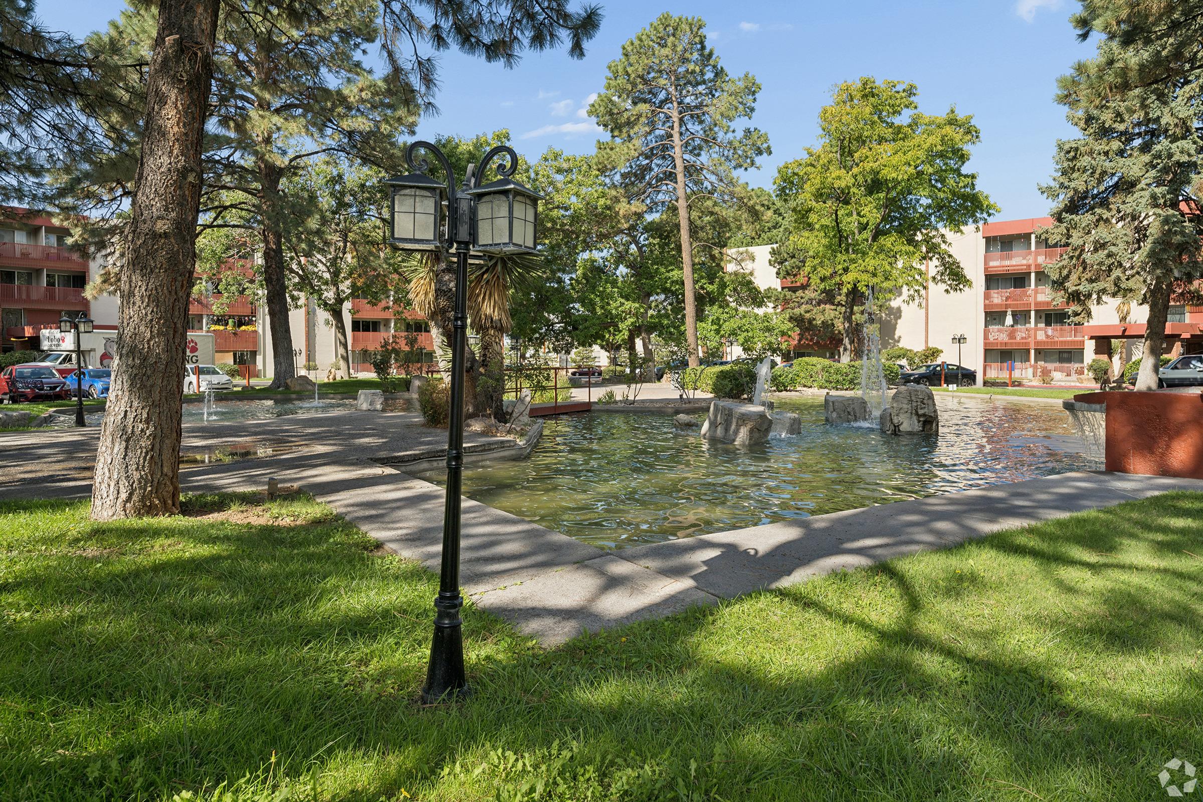 A landscaped area featuring a small pond surrounded by greenery and rocks, with a lamppost to the left. In the background, low-rise residential buildings are visible among trees under a blue sky with scattered clouds. The scene suggests a peaceful outdoor setting.