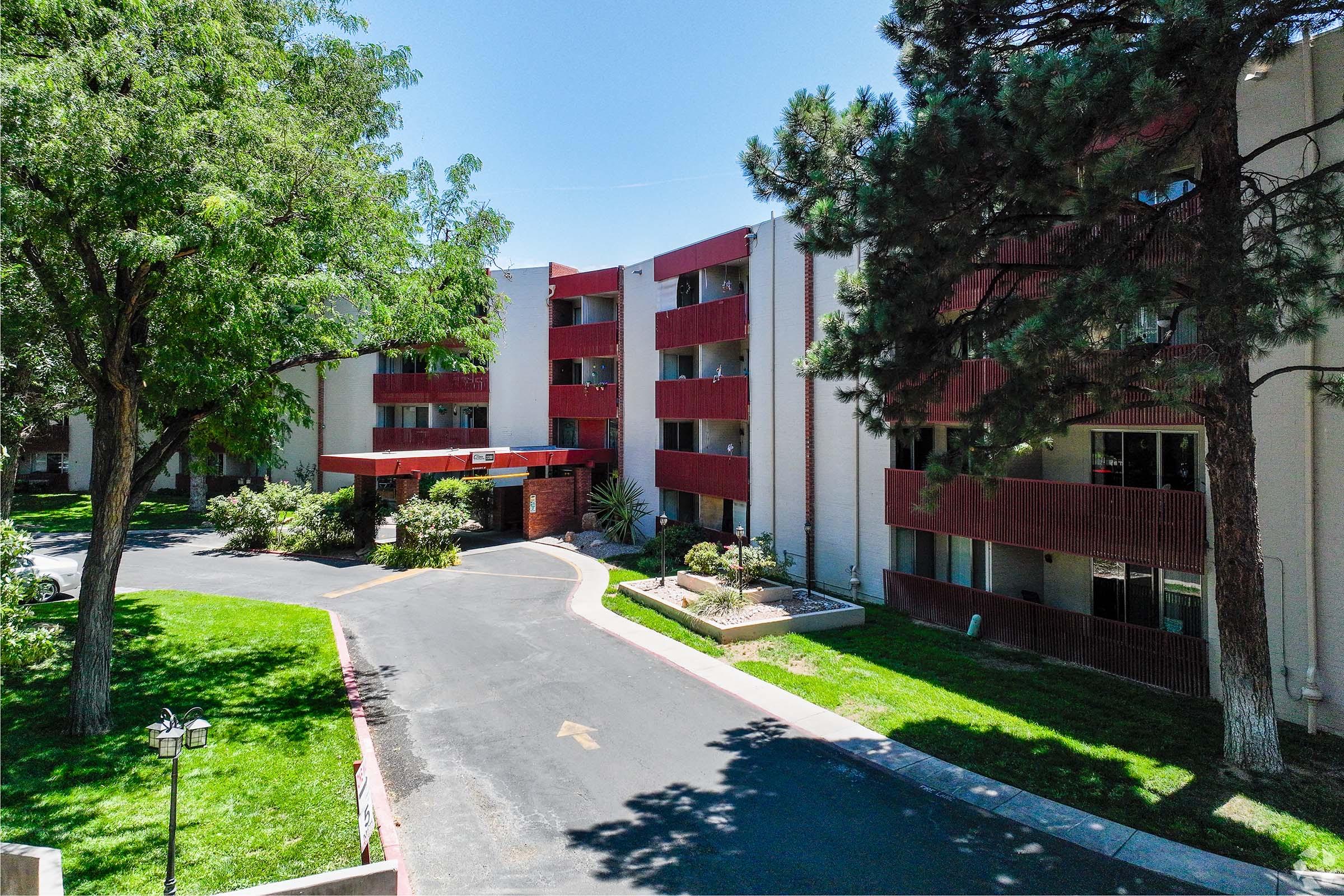 A view of a multi-story residential building surrounded by trees and well-maintained landscaping. The entrance features a pathway leading to the building, with a grassy area and flower beds enhancing the outdoor space. The sky is clear and blue, indicating a sunny day.