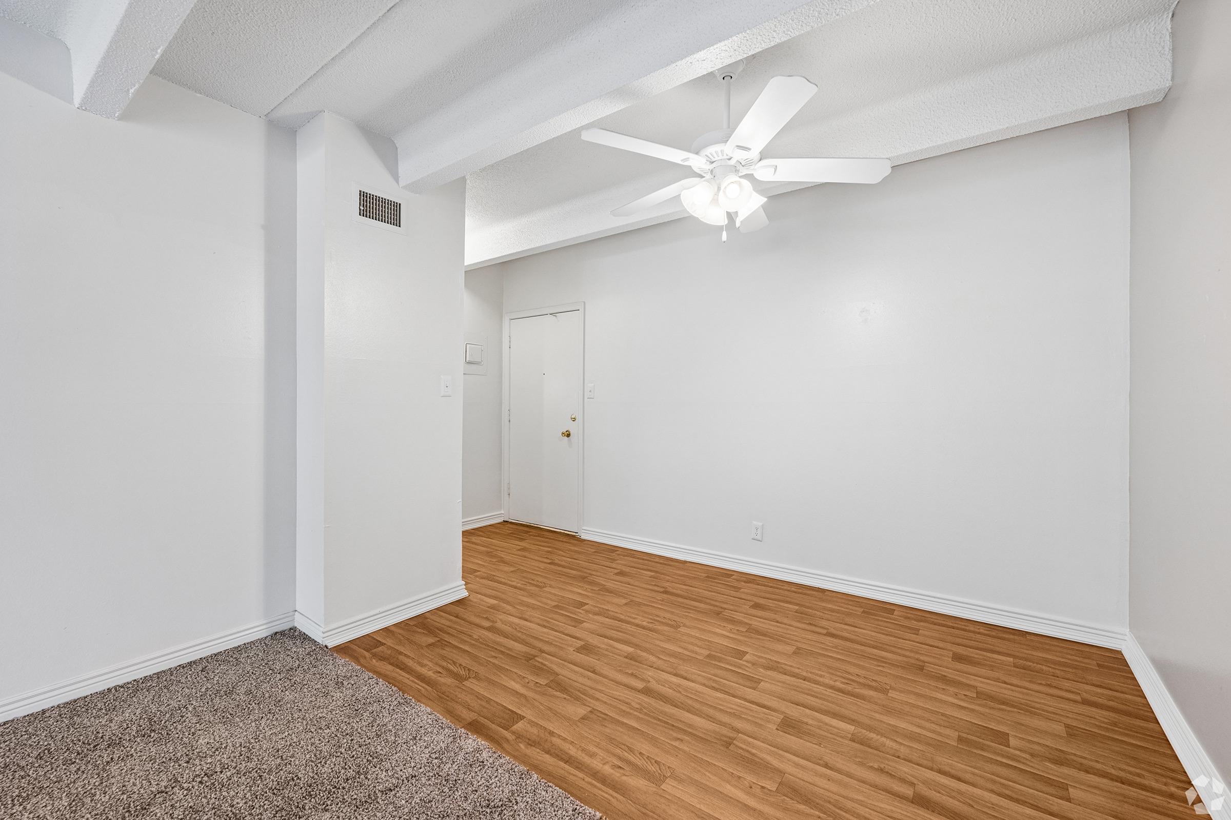 A spacious, clean living area featuring hardwood floors, a ceiling fan, and white walls. There's a doorway leading to another room, and the space is well-lit with natural light. The flooring transitions from carpet to wood, enhancing the modern feel of the room.