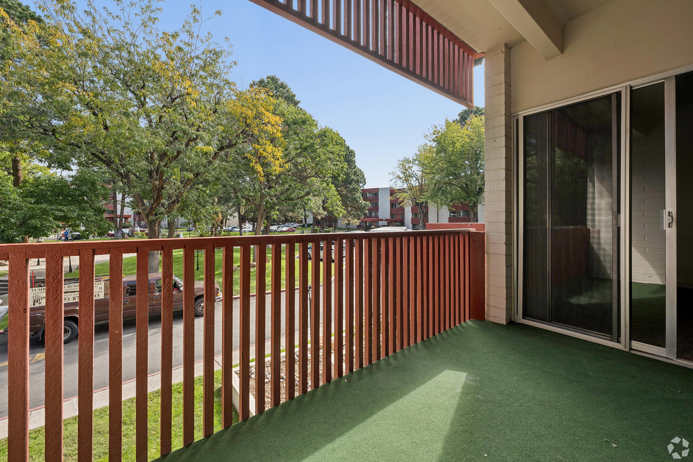 A view from a balcony featuring a green outdoor carpet, wooden railing, and large glass doors. The scene shows green trees and a landscaped area below, along with parked vehicles in the distance, creating a residential atmosphere. The sky is clear and sunny.