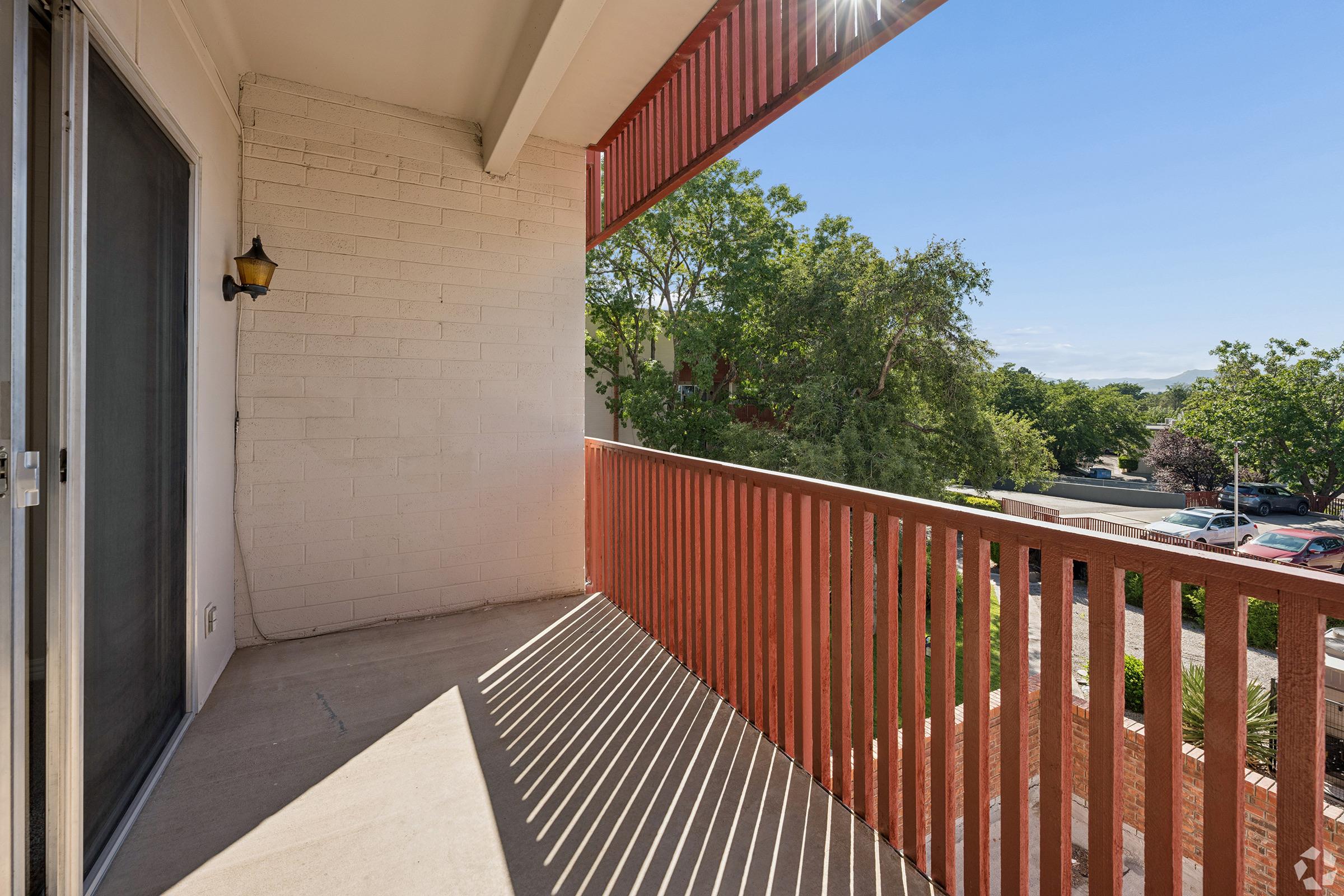 A sunny balcony with a red railing, featuring a view of greenery and a parking area. The floor is concrete, and there is a door leading indoors. Shadows cast by the railing create interesting patterns on the ground.