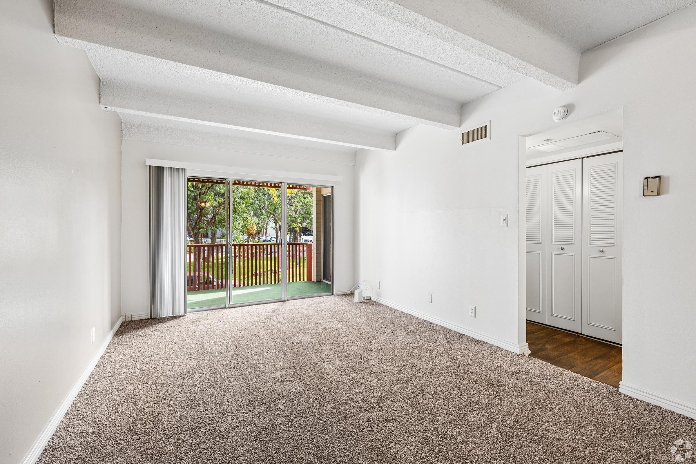 A spacious, empty living room featuring beige carpet, white walls, and exposed ceiling beams. Large sliding glass doors lead to a balcony surrounded by greenery. A closet with sliding doors is visible in the background, providing additional storage space. The room is brightly lit by natural light.