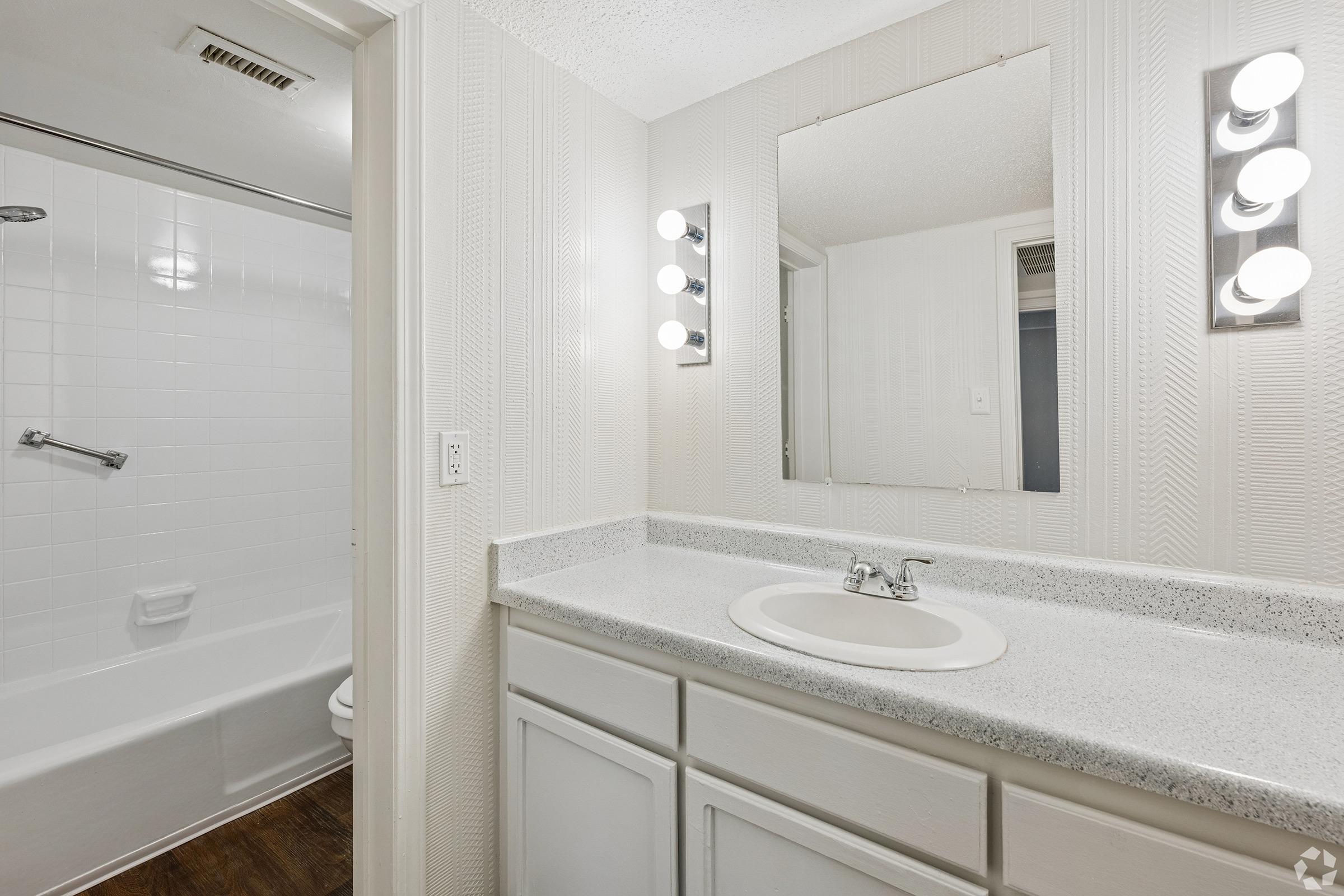 A well-lit bathroom featuring a white tiled shower with a bathtub, a light-colored countertop with a sink, and a large mirror above the sink. The walls are adorned with a textured design, and there are illuminated vanity lights. The flooring is dark wood, creating a modern aesthetic.