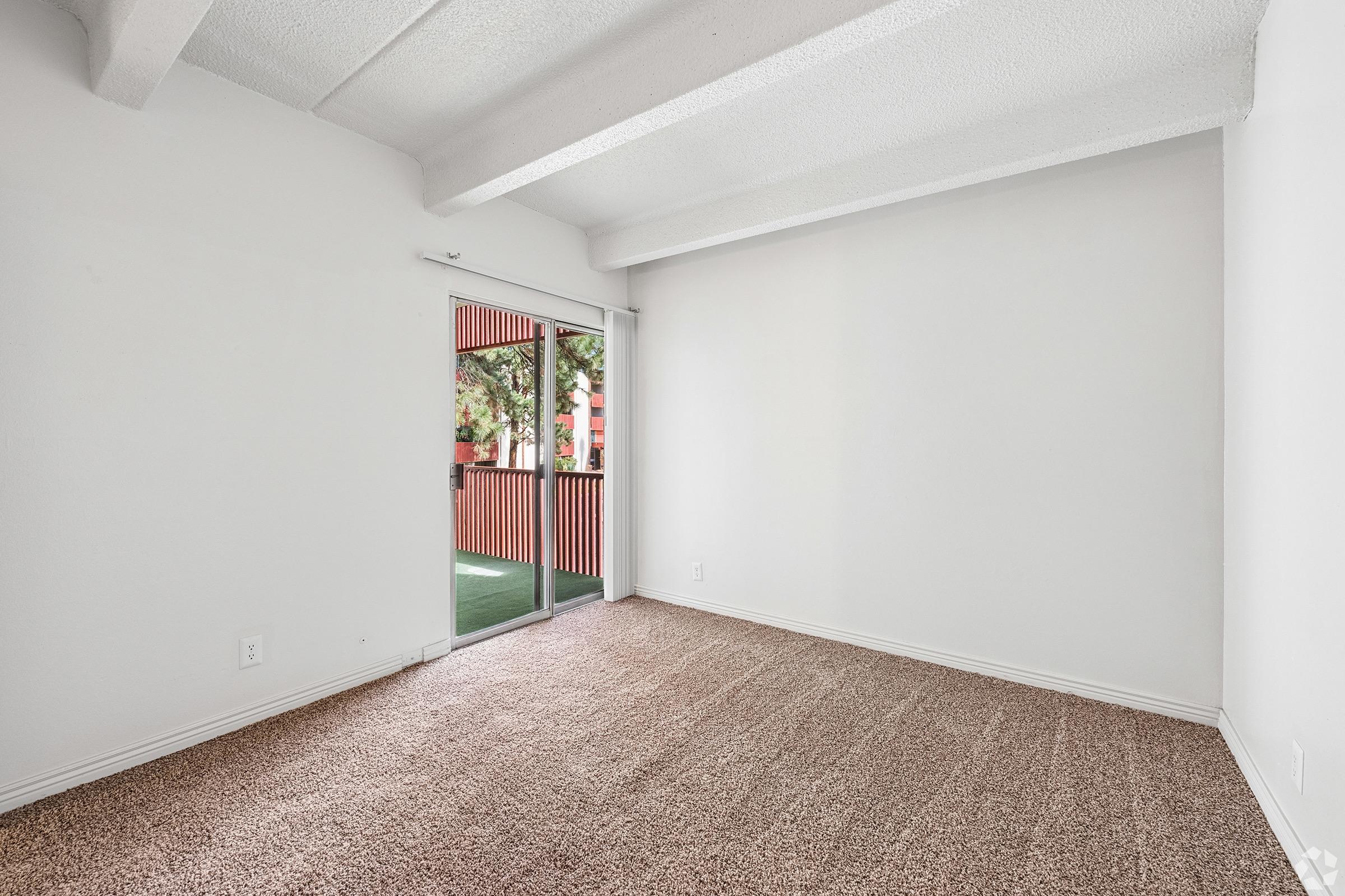A bright, empty room with light beige carpet and white walls. It features a sliding glass door that leads to a patio with a railing. The ceiling has exposed beams, and there is ample natural light coming through the door. The space is uncluttered and ready for personalization.