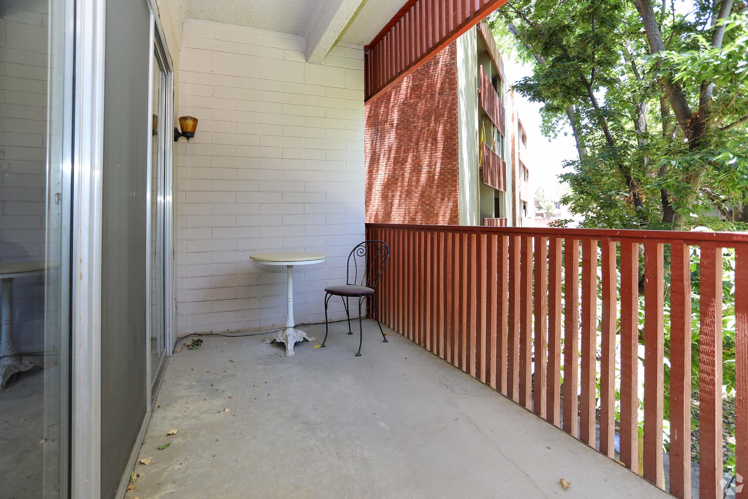 A small balcony with a round table and a single chair. It features a railing made of wooden slats, with greenery visible from the trees outside. The floor is plain and concrete, and there is a glass door leading into an interior space. Bright sunlight casts shadows on the balcony surface.