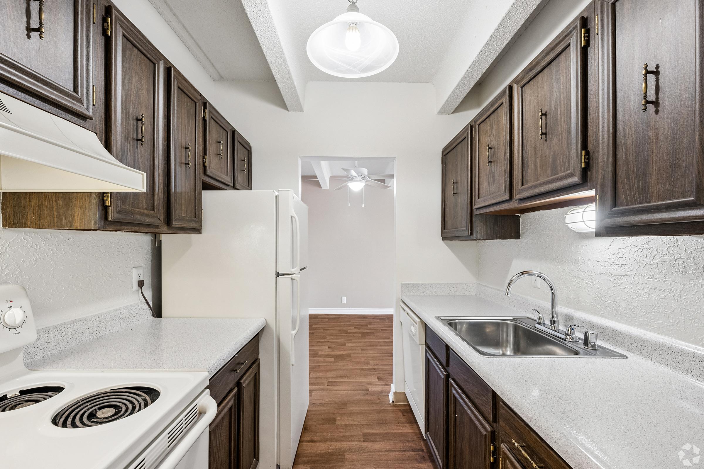 A kitchen featuring dark wooden cabinets, a white countertop, and a double sink. There is an electric stove, a refrigerator, and a ceiling light. The space has a laminate wood floor and opens into a light-colored room beyond, with visible ceiling fan and neutral walls.