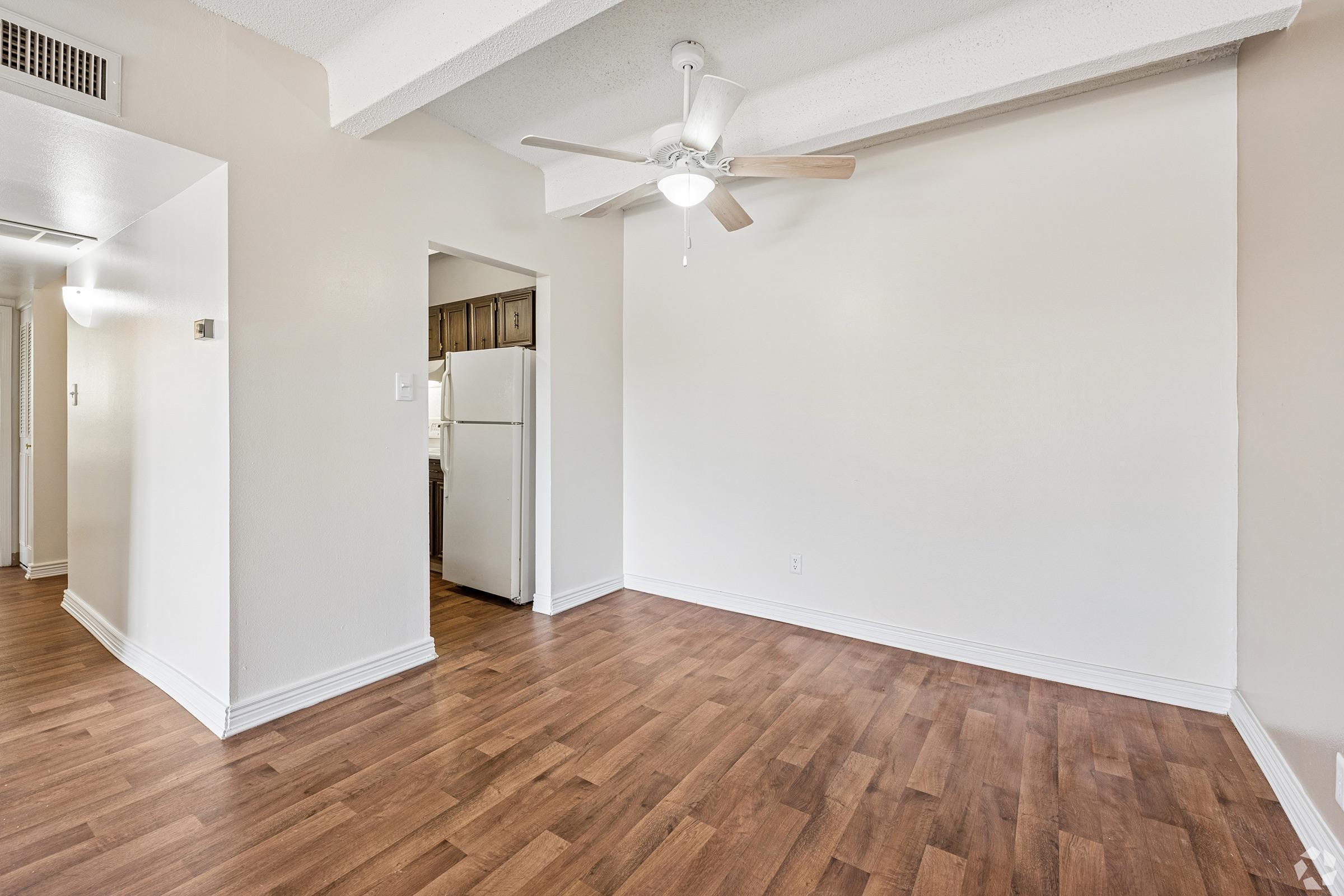 A well-lit, empty living room featuring light wood flooring, a ceiling fan, and a white wall. To the left, there's a doorway leading to a kitchen with dark cabinets. The room has a simple, clean design with ample space for furniture, creating a cozy atmosphere.